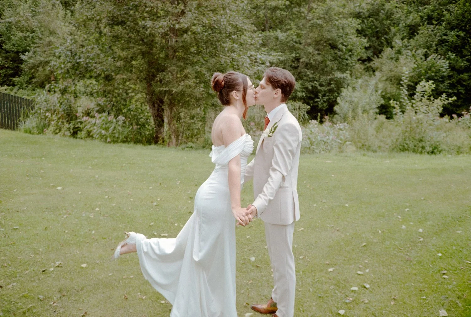 A bride and groom holding hands and sharing a kiss outdoors on a grassy area with trees in the background.