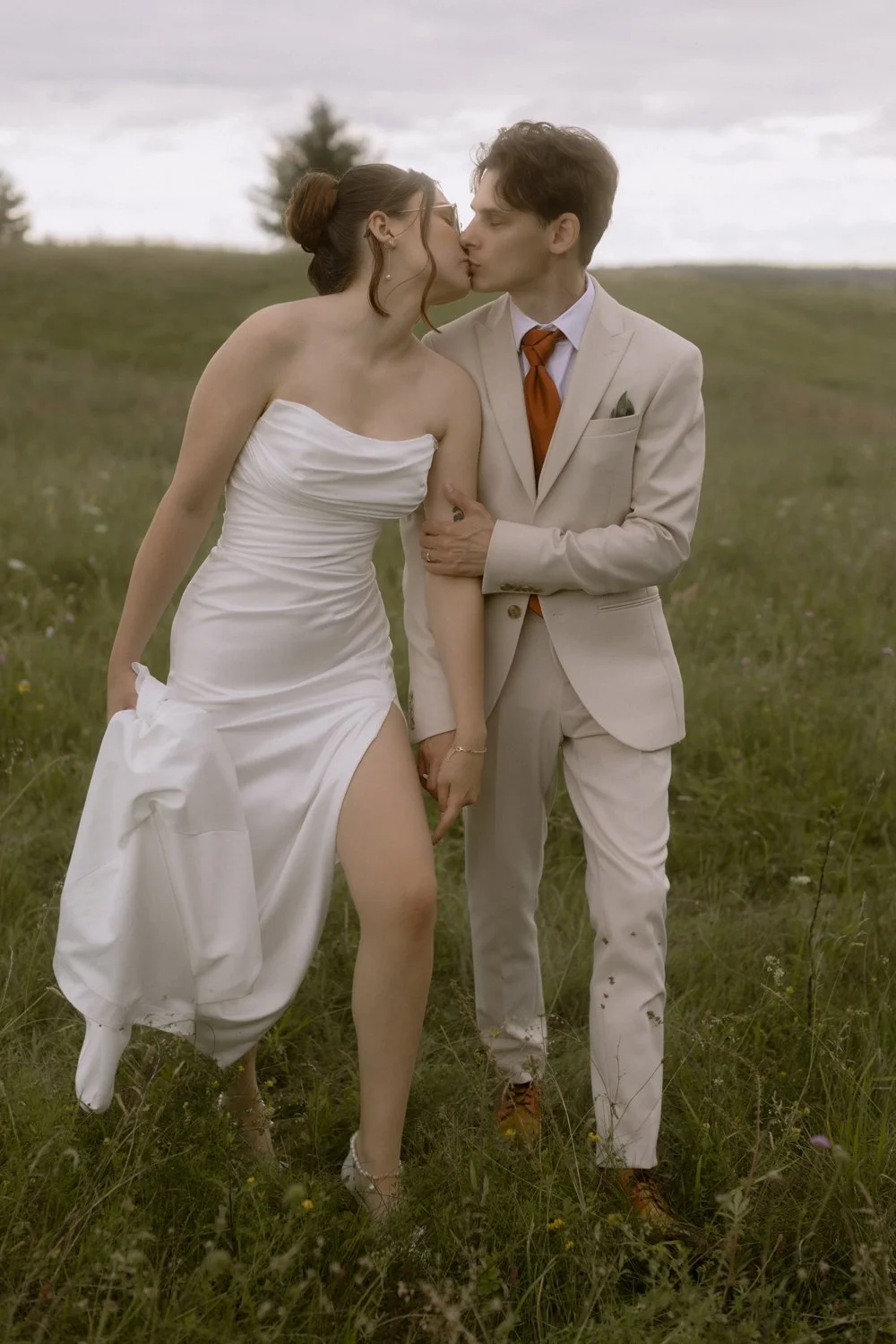 A couple dressed in wedding attire sharing a kiss outdoors in a grassy field, with the woman in a strapless white gown and the man in a cream-colored suit with a brown tie.