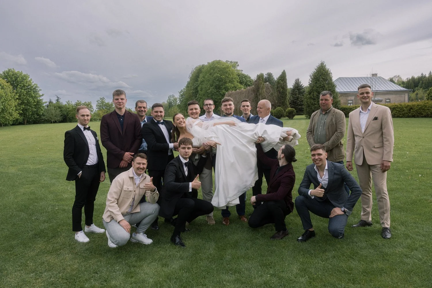 A group of people at a wedding, with the bride in a white dress being carried horizontally by friends, outdoors on a grassy area with trees and a house in the background.