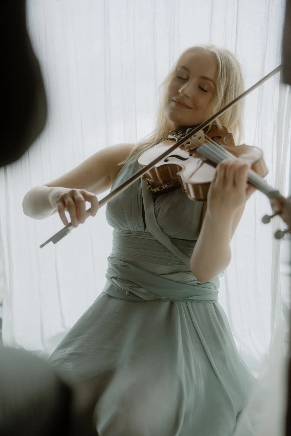 A woman in a light gray dress playing the violin with closed eyes, standing in front of sheer curtains.