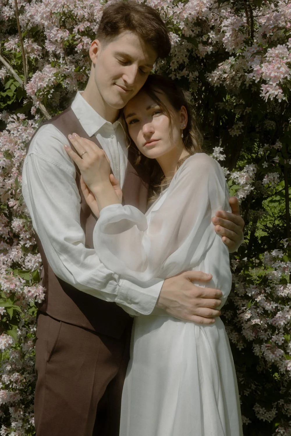 A young couple embracing in front of blooming pink and white flowers on a sunny day.