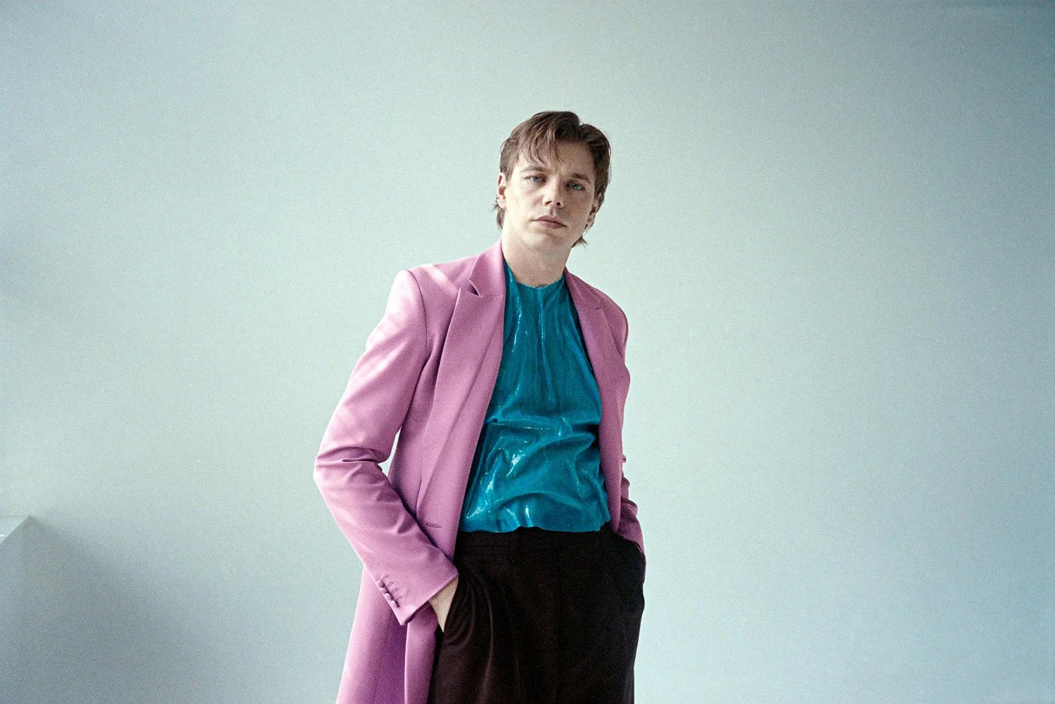 A young man with styled brown hair wearing a pink coat over a shiny blue shirt and black pants, standing against a plain light-colored background.