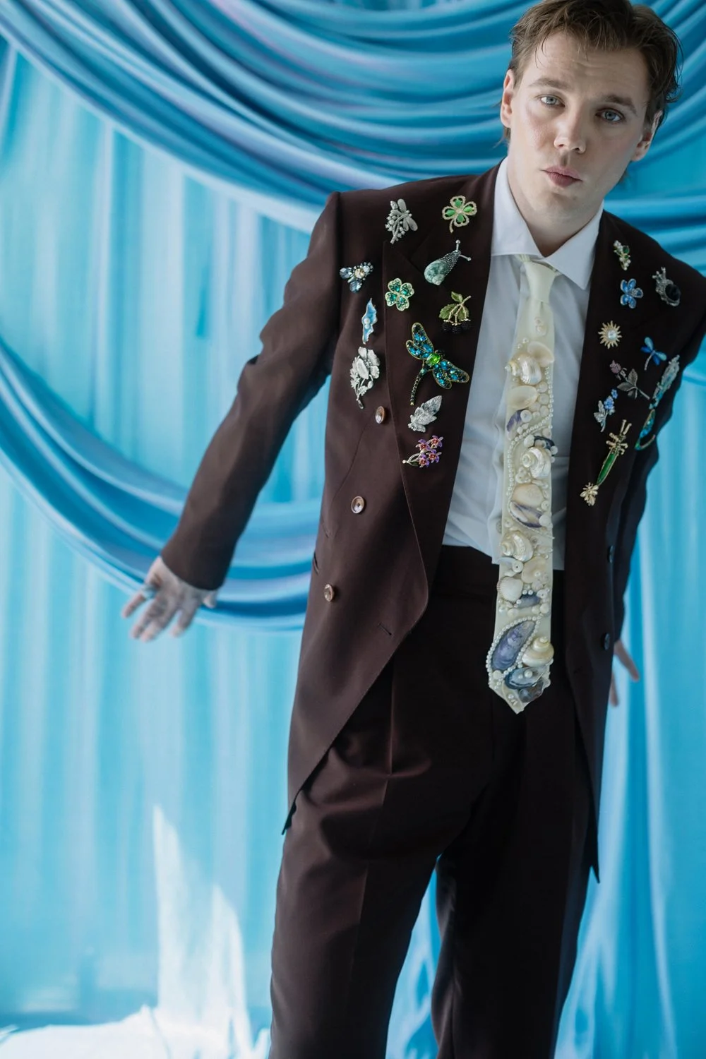 A young man in a brown suit with various decorative pins and brooches, standing in front of a blue, draped fabric backdrop.