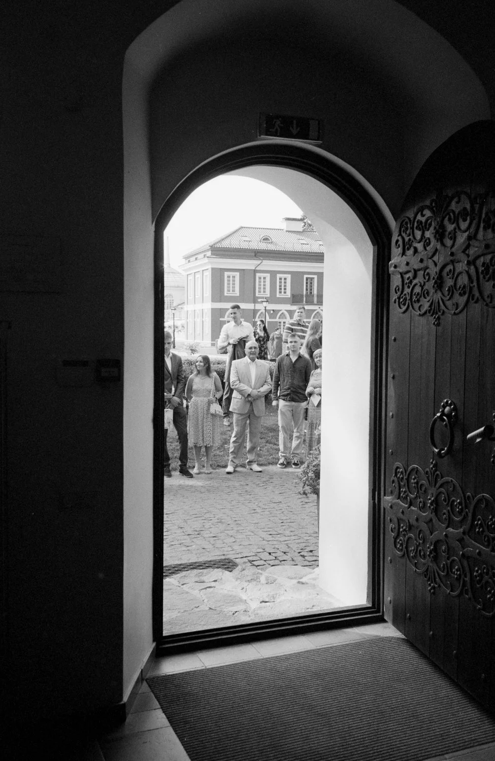 Group of people gathered outside, seen through the open door of a building, with some individuals standing on the cobblestone pathway, and a large building in the background.