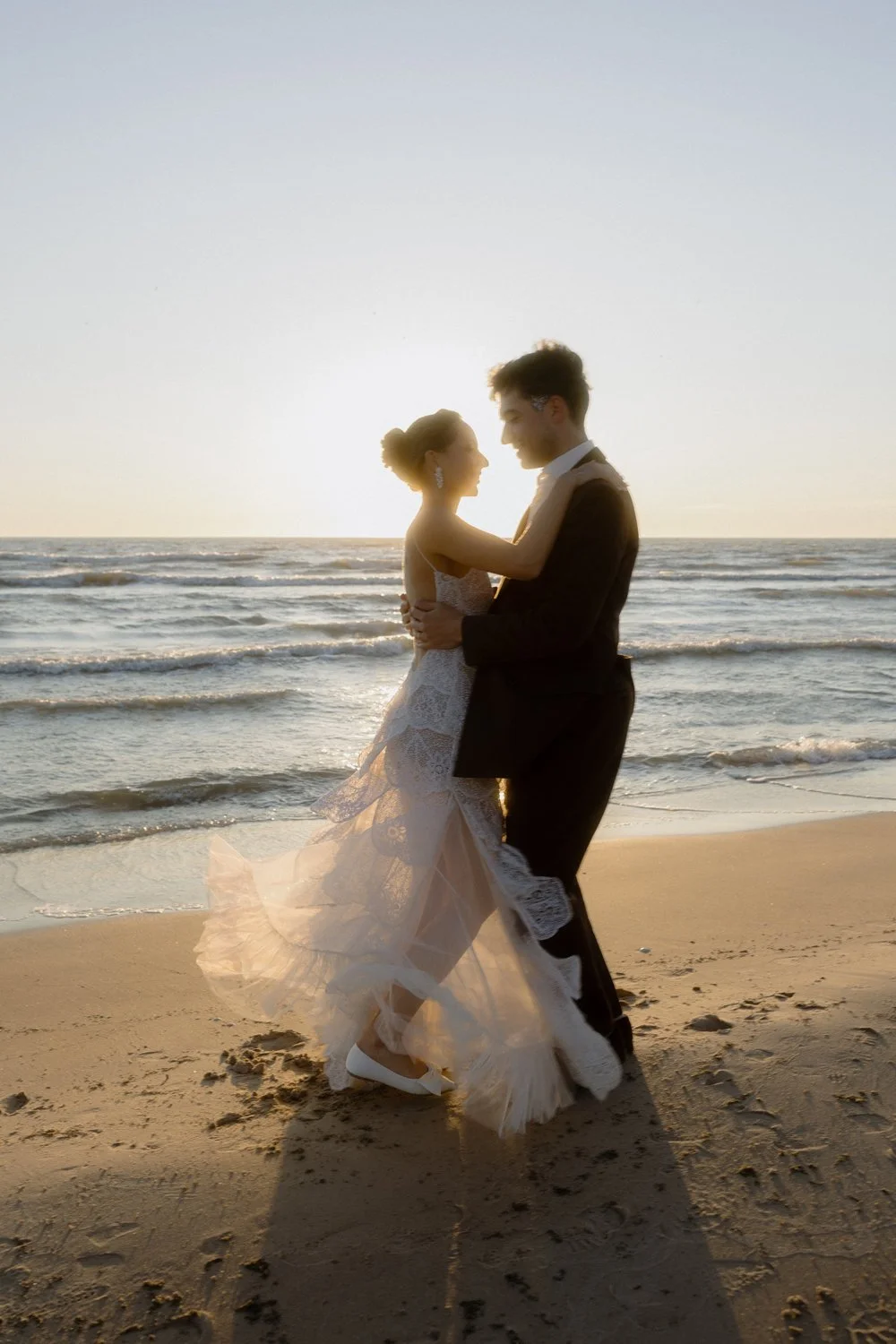 A couple in wedding attire dancing on the beach at sunset.