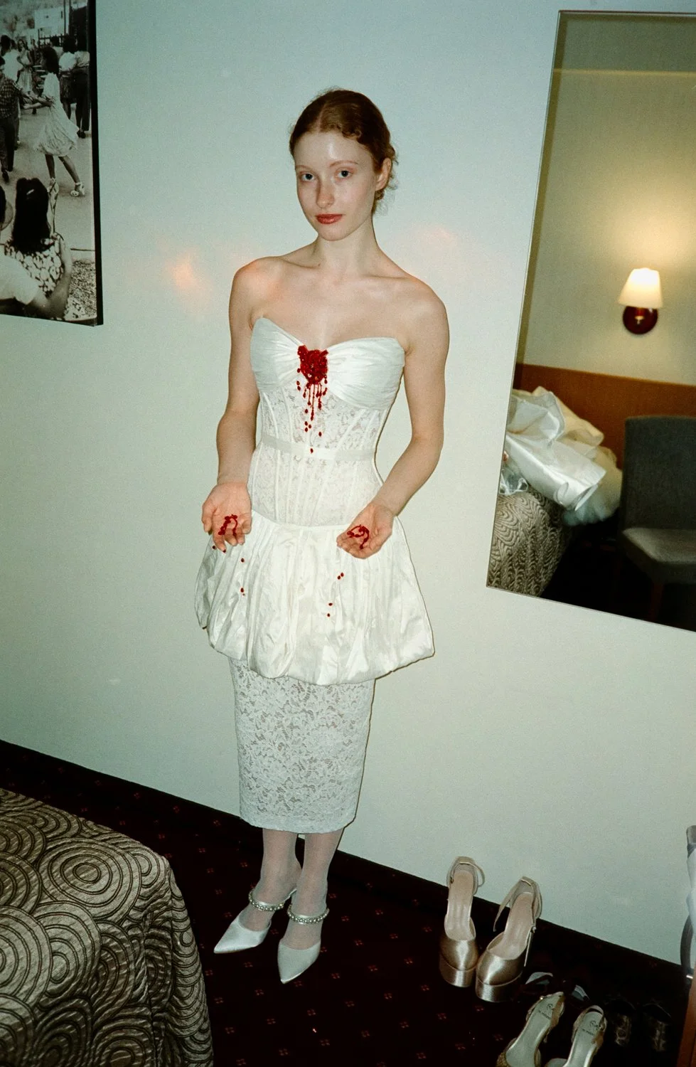 A woman wearing a strapless white wedding dress with red details, standing in a room with a mirror and a bed in the background, and several pairs of shoes on the floor.