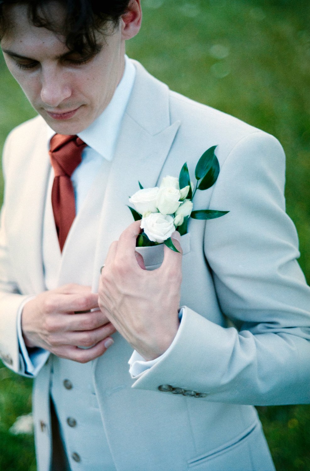 A person dressed in a white suit with a rust-colored tie holding a white boutonniere on their lapel, standing outdoors on a grassy background.