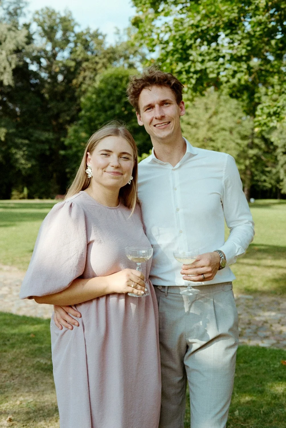 A smiling woman and man stand outdoors in a park, holding glasses of white wine, with green trees and grass in the background.