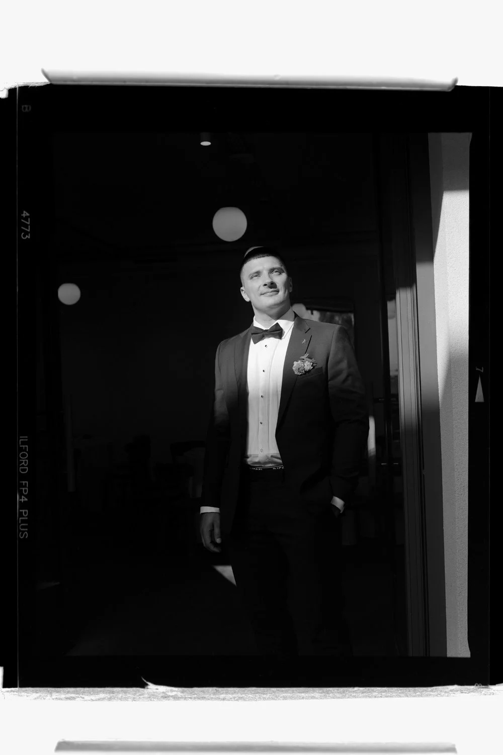 A man in a tuxedo and bow tie, standing in a doorway, looking off into the distance in sunlight, black and white photograph.