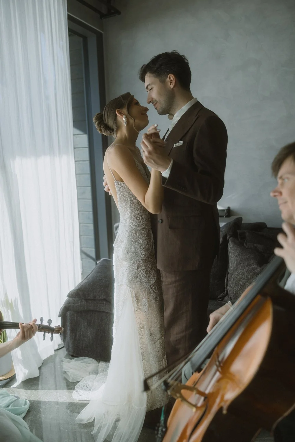A bride and groom are dancing closely indoors with sunlight streaming through sheer curtains. The bride is in a lace wedding dress, and the groom is in a dark suit. There is a musician playing a cello nearby, and a person playing a guitar.
