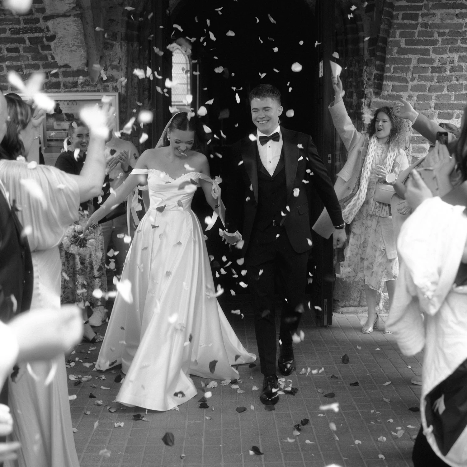 A newlywed couple walking hand in hand through a shower of flower petals, smiling, with friends and family celebrating around them outside a brick building.