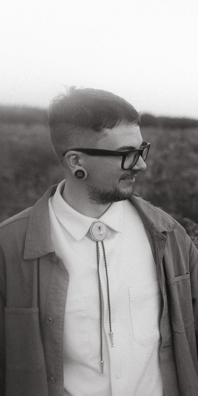 A black and white photo of a man with short hair, glasses, and ear gauges, smiling and looking to his left in an outdoor setting.