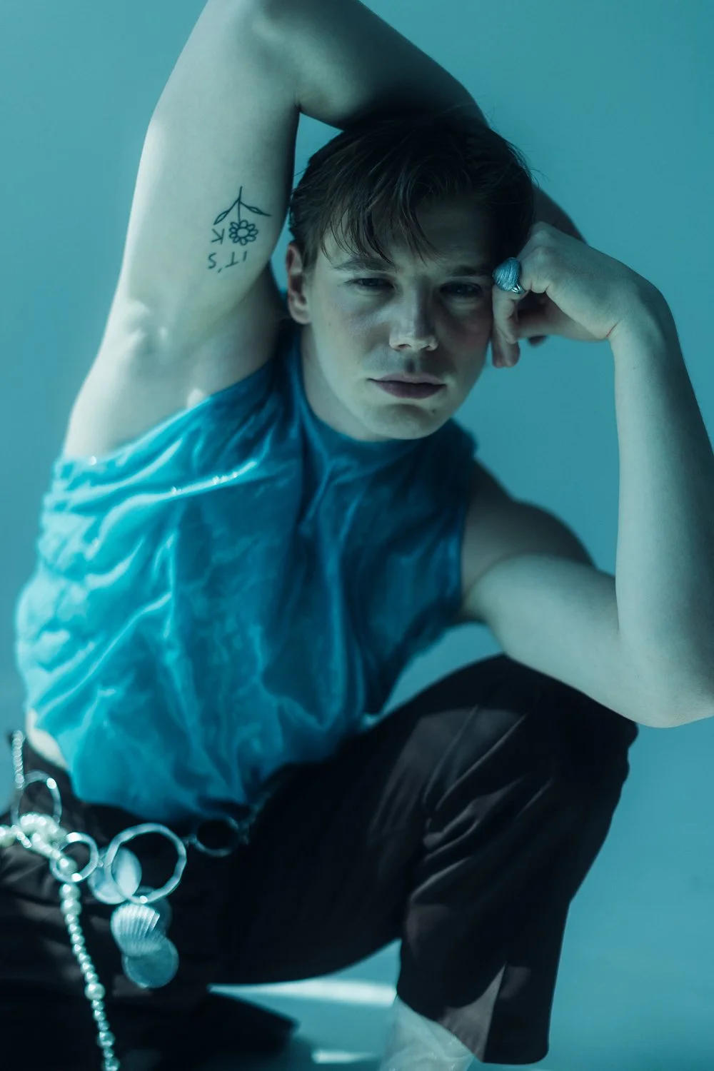A young man with short, styled hair, wearing a blue sleeveless shirt and black pants, seated with one knee up. He leans his head on his hand, looking into the camera with a serious expression. He has a tattoo on his bicep and is wearing a large ring 