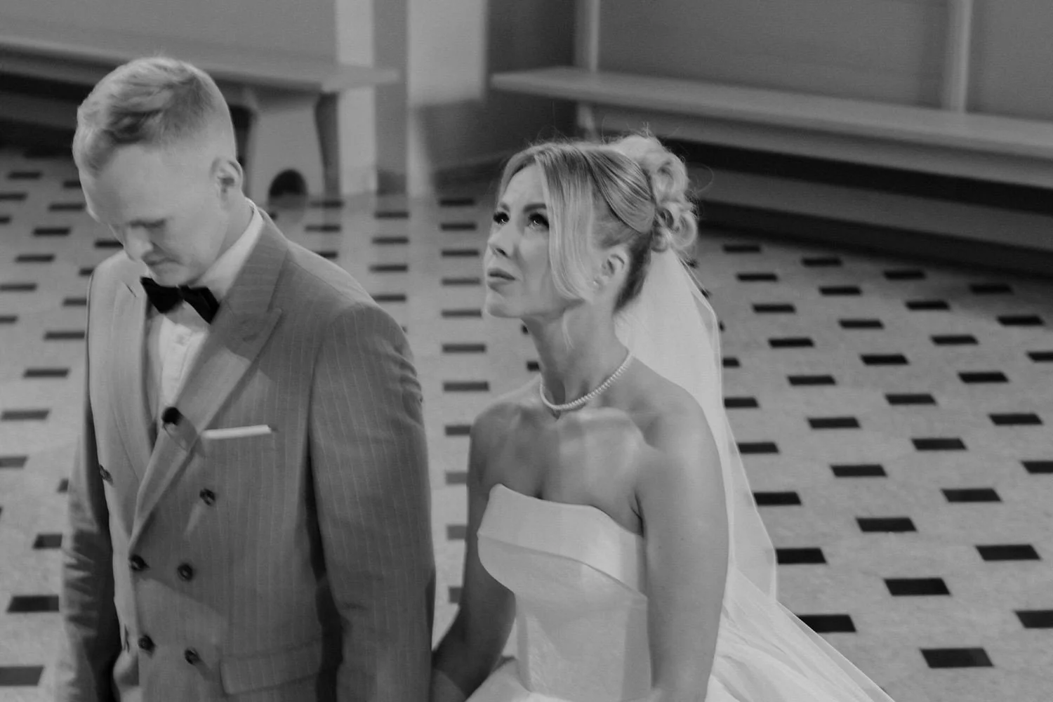 A black-and-white photo of a bride and groom at a wedding, with the bride looking upward and holding her wedding dress, and the groom looking downward, in a formal indoor setting.
