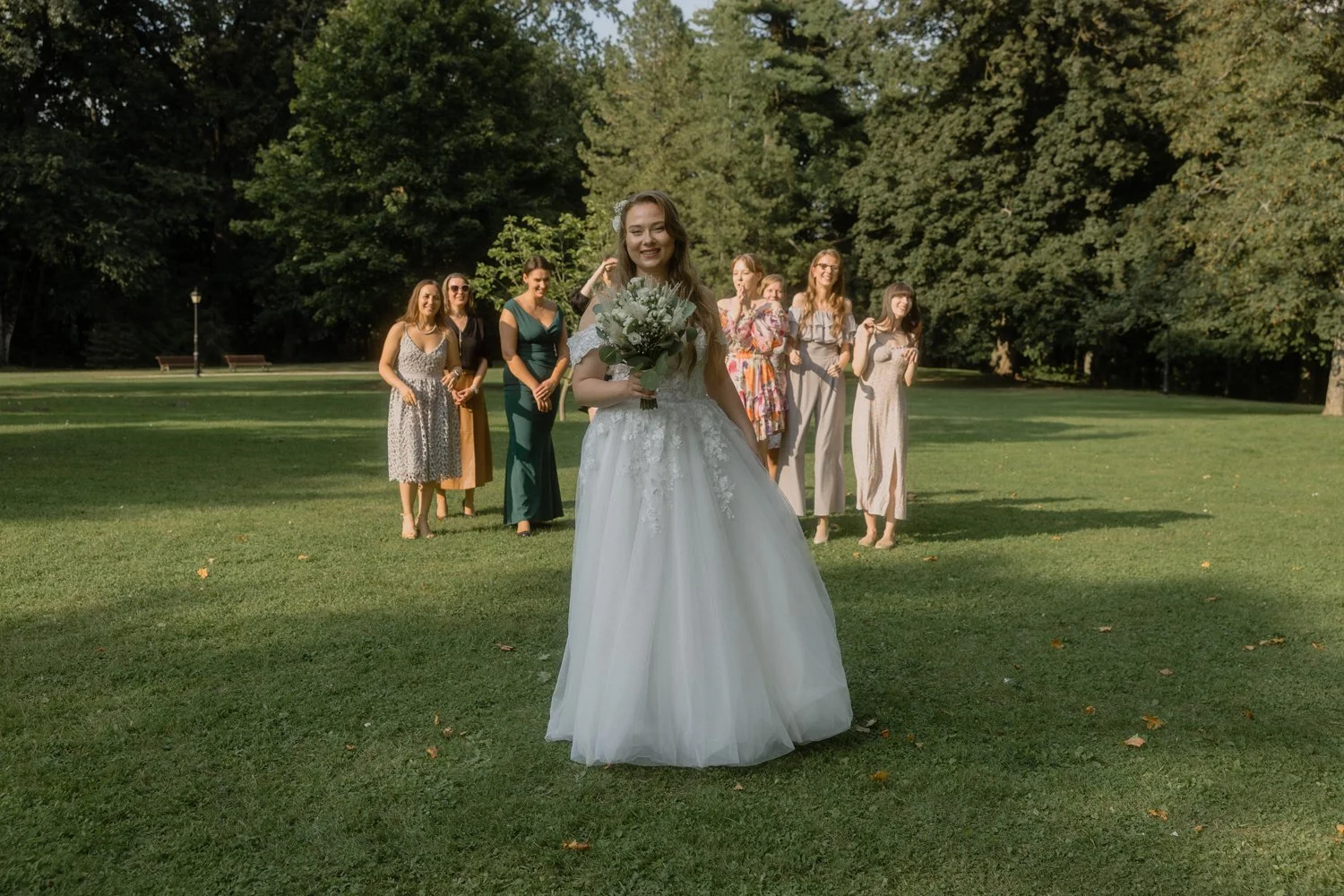 A bride in a white wedding dress holding a bouquet of flowers, standing on a grassy field with her bridesmaids in colorful dresses in the background, surrounded by trees.