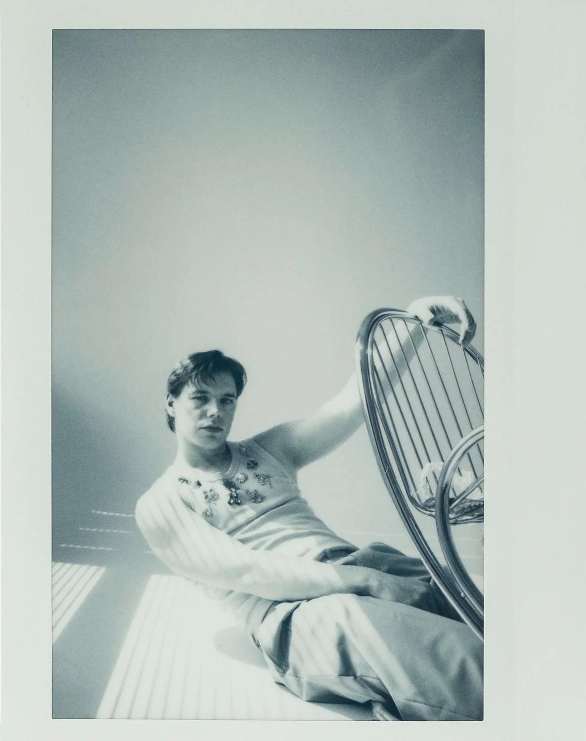 Black and white photograph of a young man sitting on a stripy lounge chair, looking at the camera, with his arm resting on a matching chair, against a plain wall.