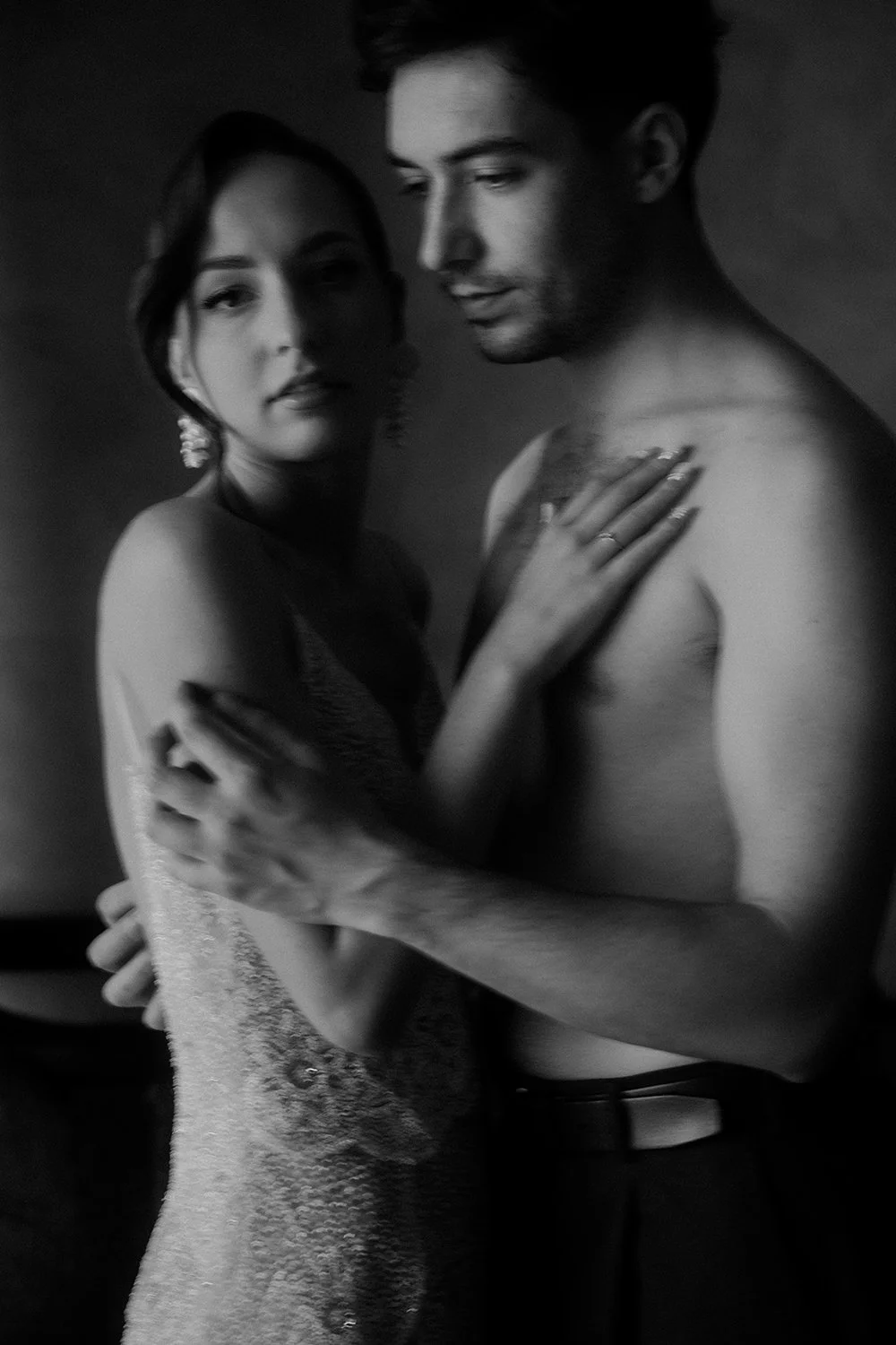 A black and white photo of a man and woman embracing, both shirtless, with the woman wearing a strapless dress and earrings, and the man wearing a belt. They are looking at the camera with serious expressions.