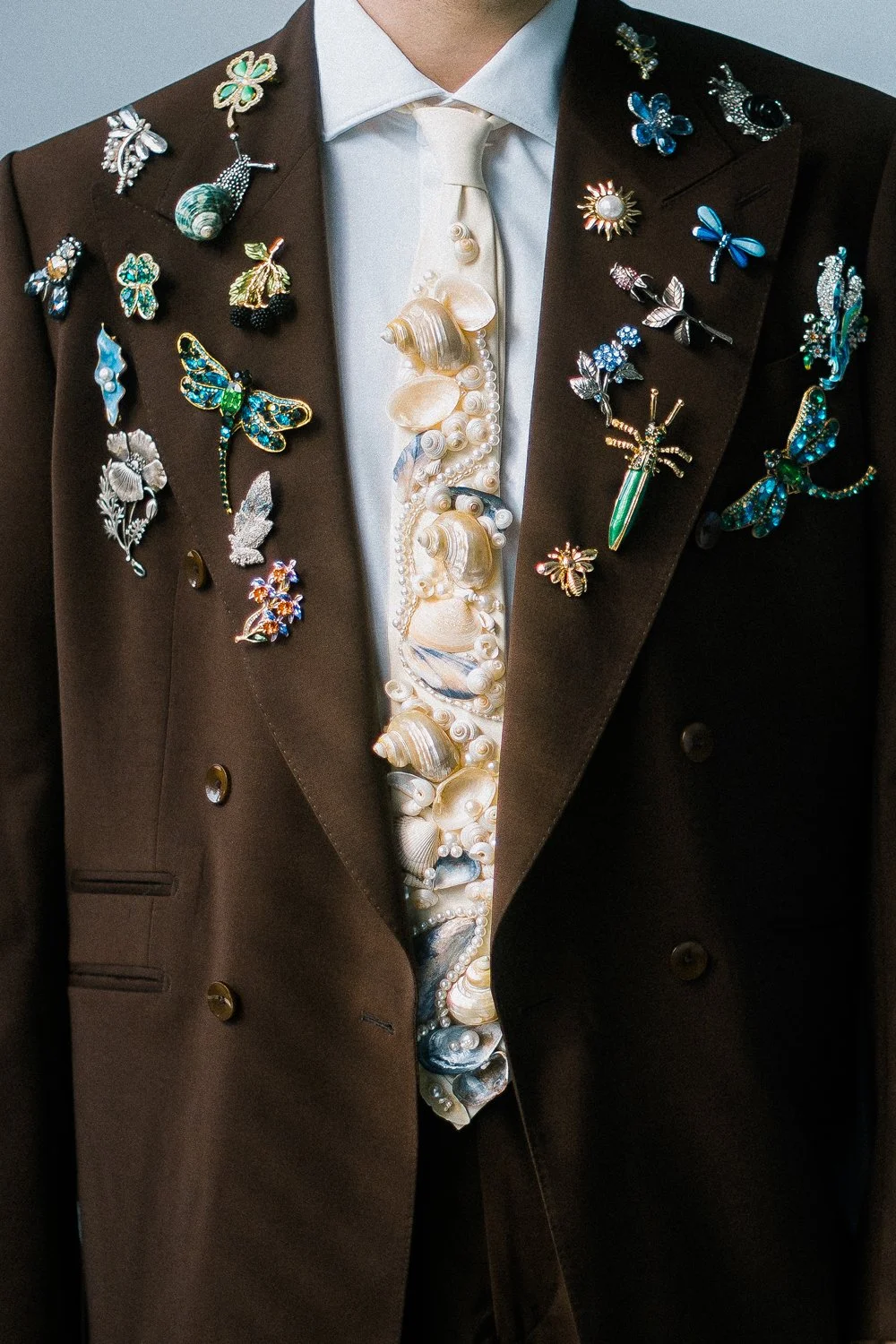 A person wearing a dark brown double-breasted blazer decorated with colorful, intricate insect and nature-themed pins, a white shirt, and a cream-colored necktie with shells and pearls.