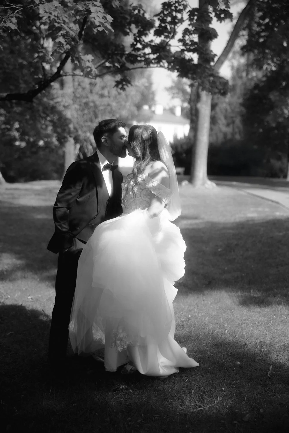 A black and white photograph of a bride and groom sharing a kiss outdoors. The groom is dressed in a suit with a bow tie, and the bride is wearing a white wedding gown with a veil. They are surrounded by trees and the sunlight filters through the lea