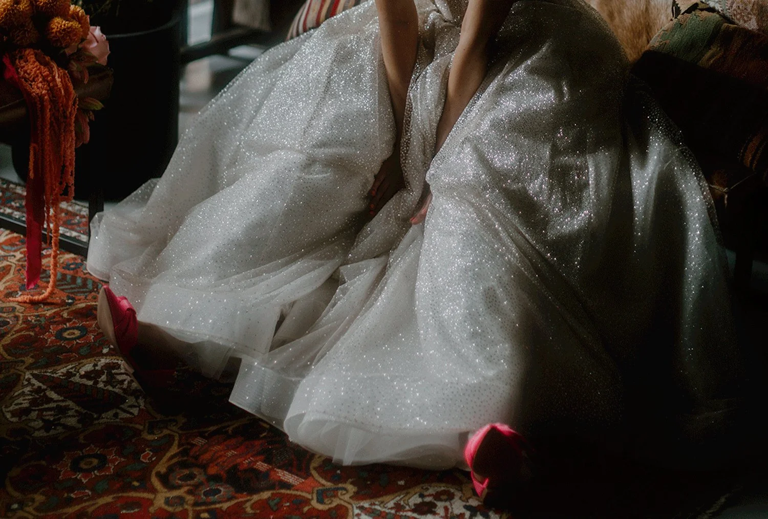 A bride wearing a sparkling, light-colored gown and red high heels is sitting on the floor, with their hands resting on their lap. The scene includes a patterned rug and some furniture in the background.