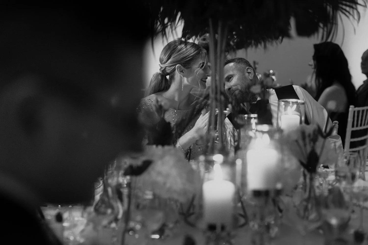 Black and white photo of a bride and groom seated at a wedding reception table, smiling and leaning close to each other, surrounded by candles and floral decorations.