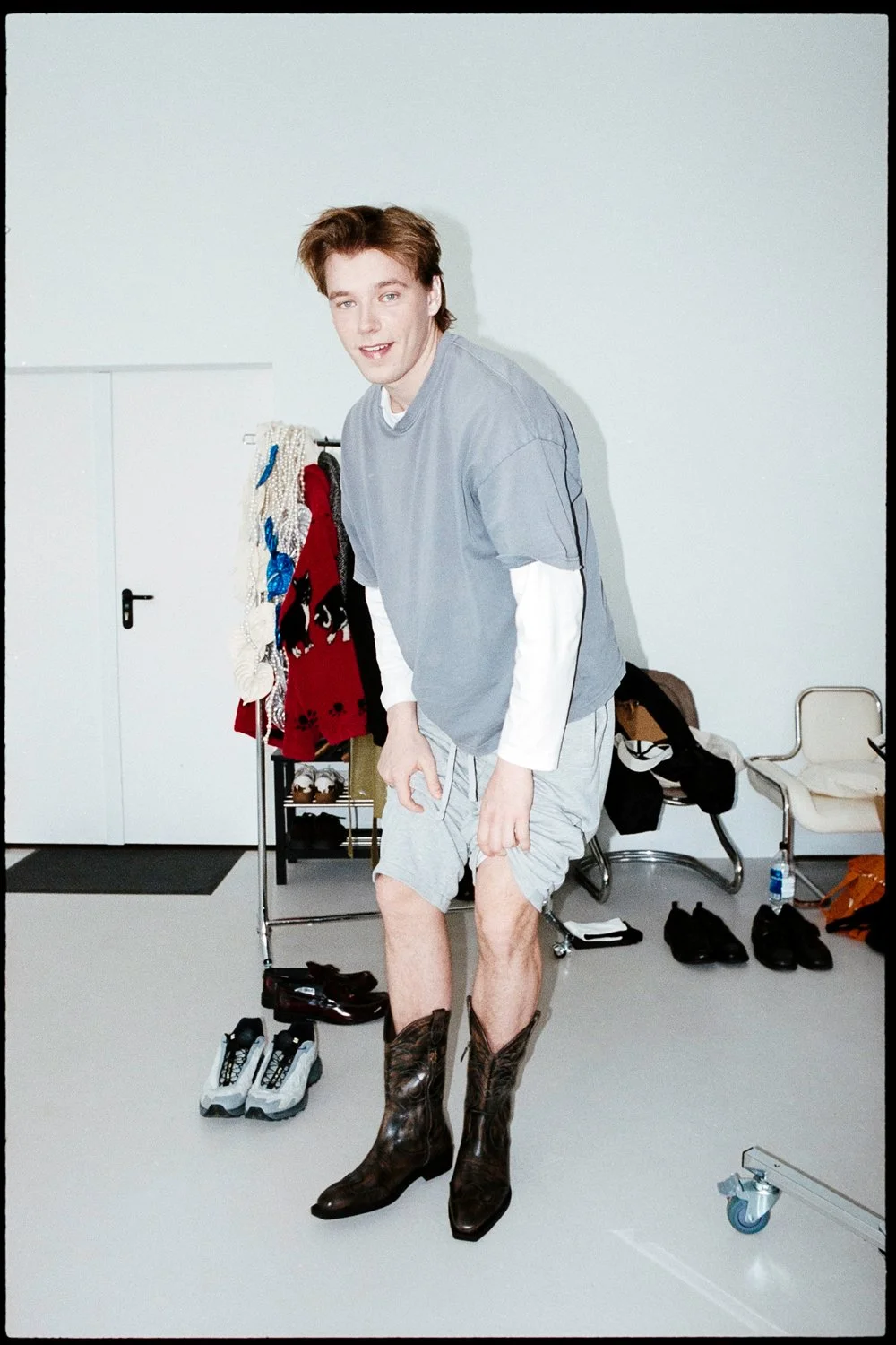 A man standing in a room, wearing a white long-sleeve shirt, a grey t-shirt layered over it, grey shorts, and brown cowboy boots. The room has various clothing items and shoes scattered around, with a clothing rack and a white chair against the wall.