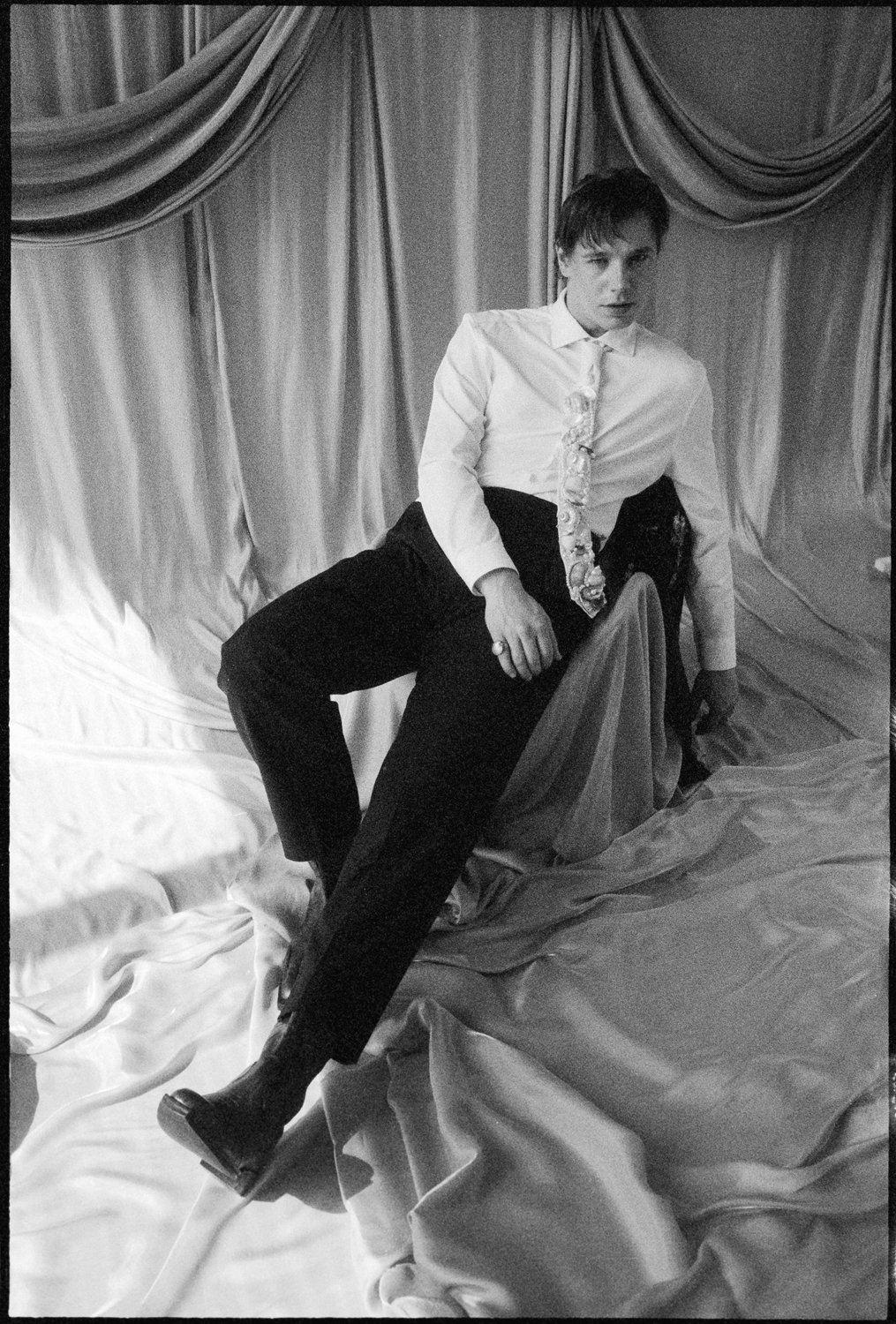 A person wearing a white shirt, tie, and dark pants, posing against draped fabric and bed linens in a black and white photo.