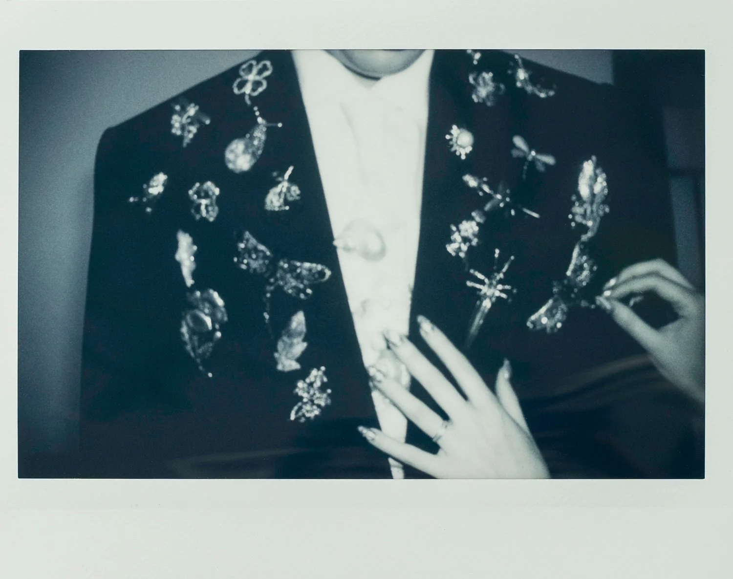 Black and white photo of a person wearing a jacket decorated with various shiny pins and brooches, with a hand resting on their chest showing rings on fingers.