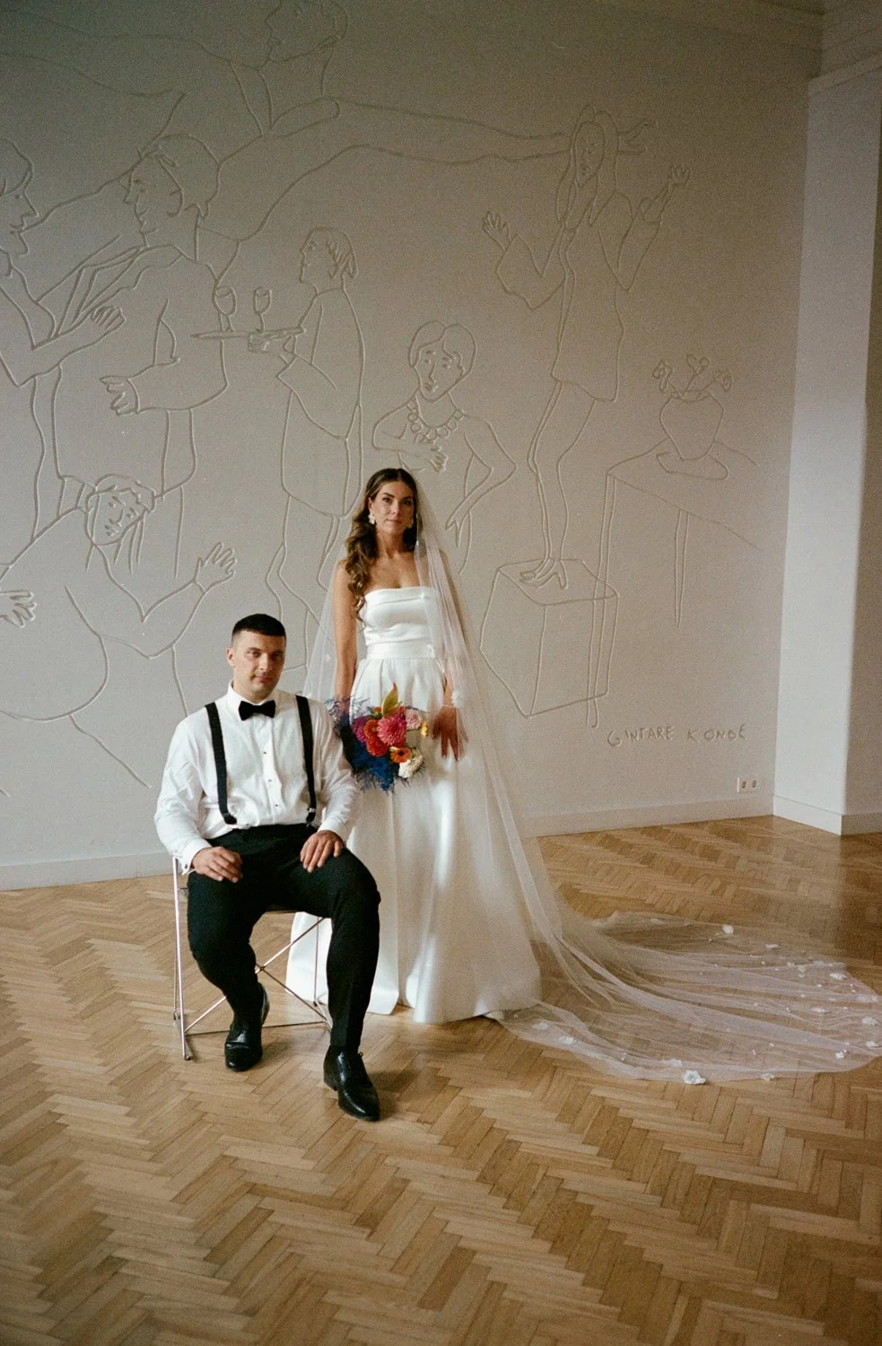 A bride in a white wedding gown holding a bouquet standing next to a seated groom in a tuxedo with suspenders. They are indoors in front of a wall with line art drawings of people and objects.