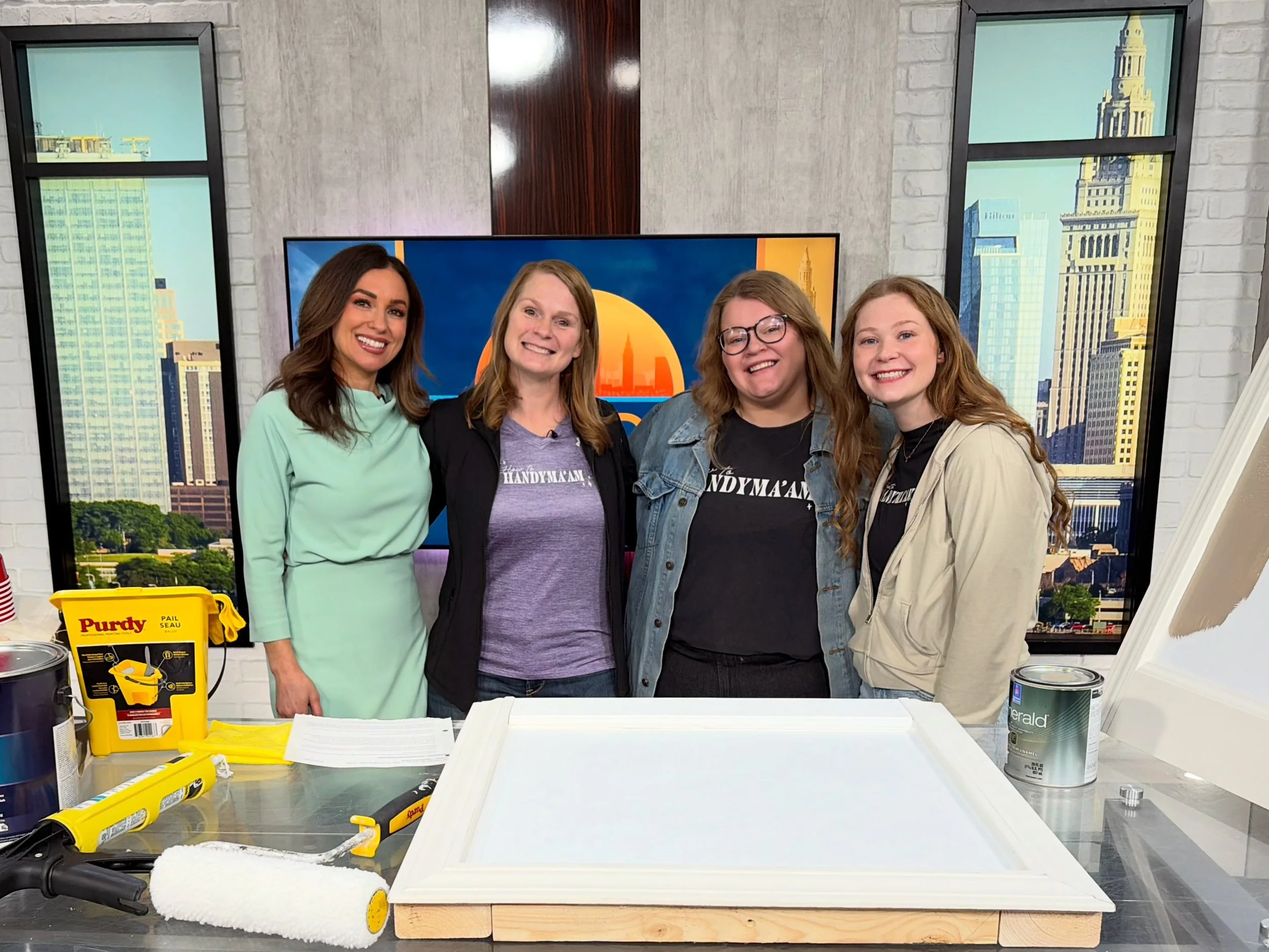 Four women standing behind a table with painting supplies, including paint cans, a paint roller, and a yellow paint tray, in a room with large window high-rise views of buildings.