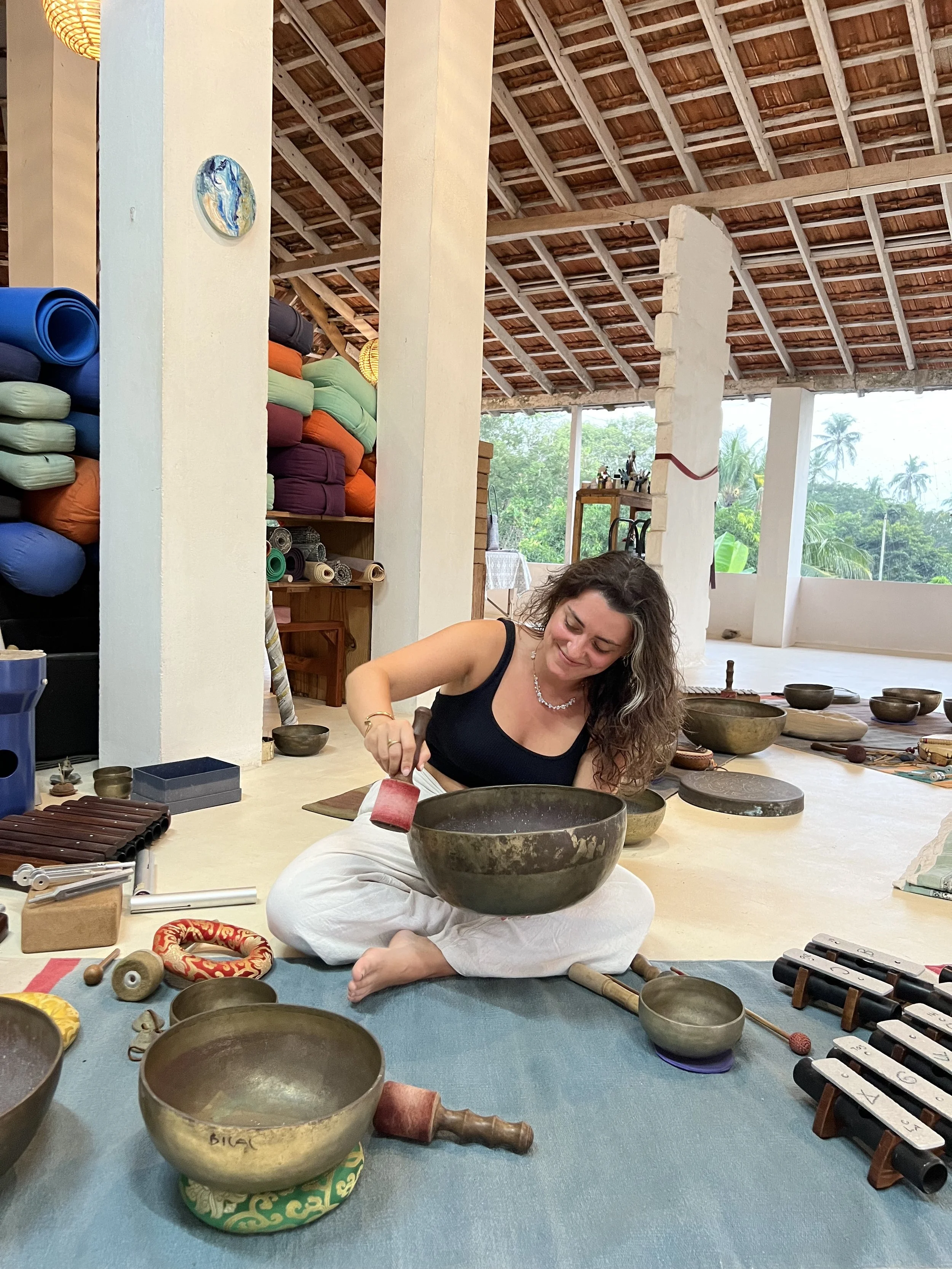 Woman sitting cross-legged on the floor, playing singing bowls in a spacious room with a high wooden ceiling and large windows showing green trees outside. The room has various musical instruments and colorful stacked yoga mats against a wall.