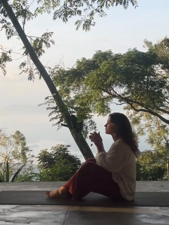 A woman sitting cross-legged on a yoga mat outdoors, practicing meditation or prayer with her hands clasped together, surrounded by trees and nature.