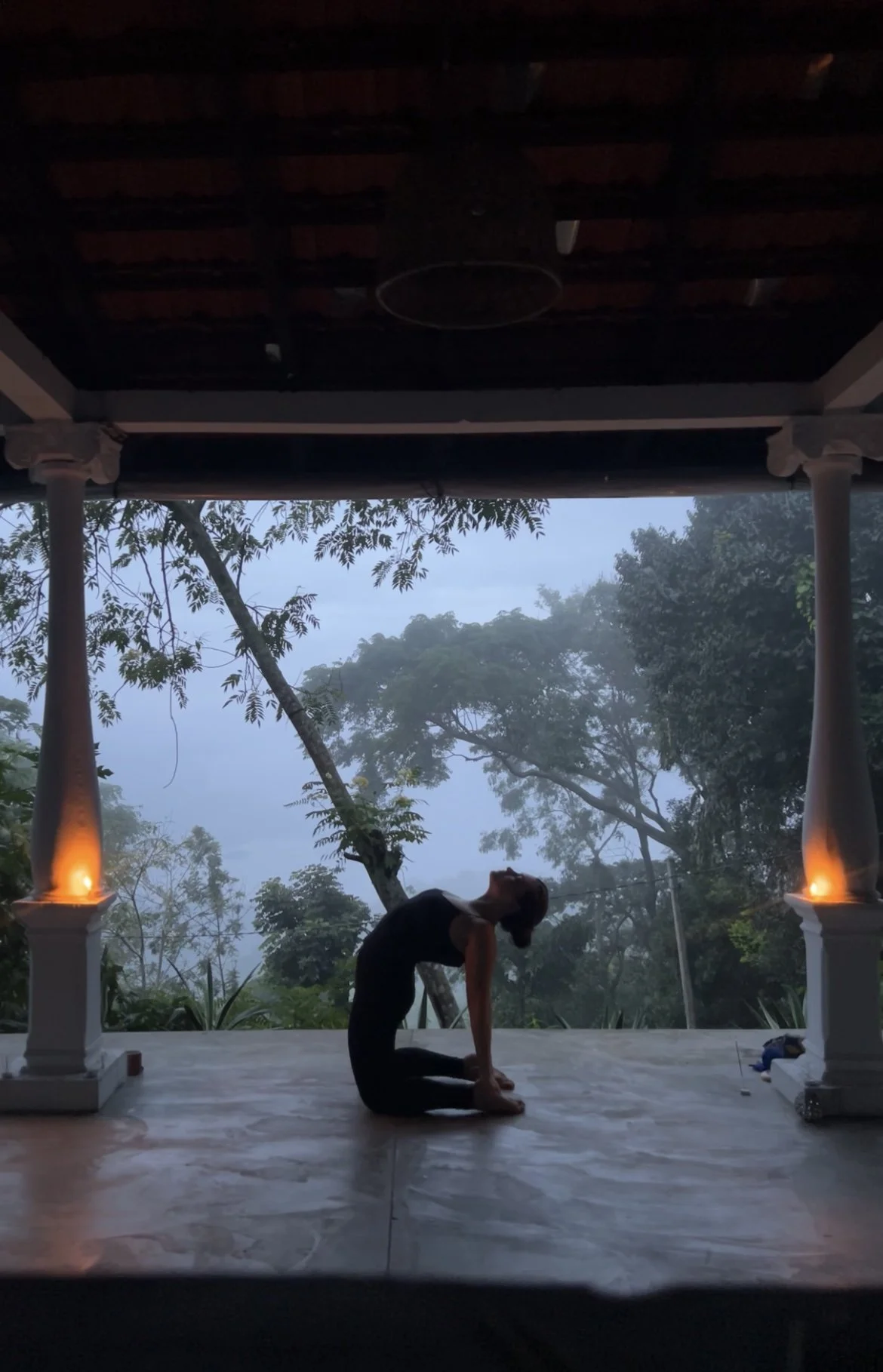 A person practicing yoga outdoors during dawn or dusk on a deck with white columns, surrounded by trees and mist, with two lit torches on either side.