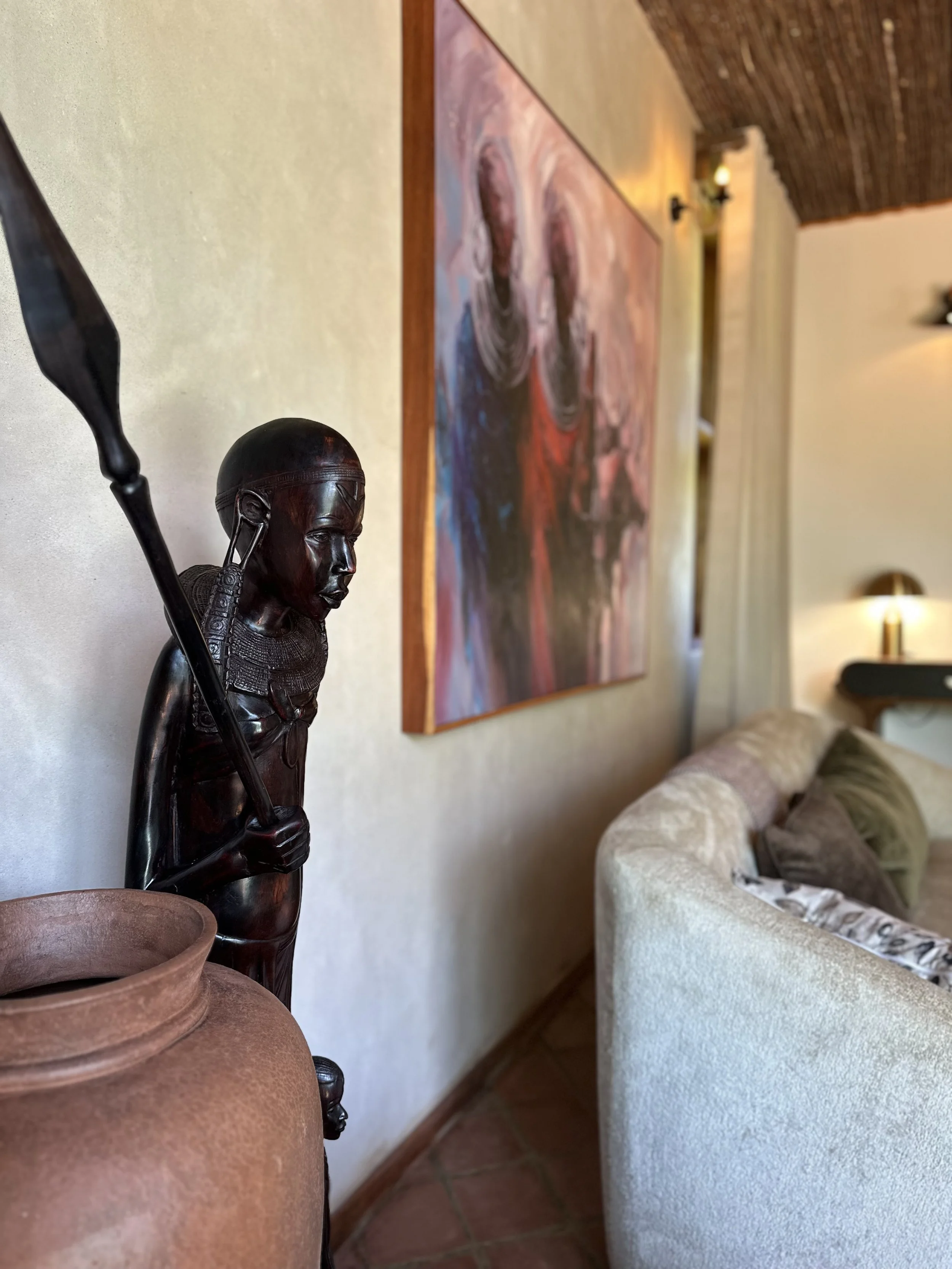 A dark wooden statue of a young indigenous person holding a spear, placed near a large clay pot in a living room. In the background, there is a colorful abstract painting on the wall and a cozy beige couch with pillows.