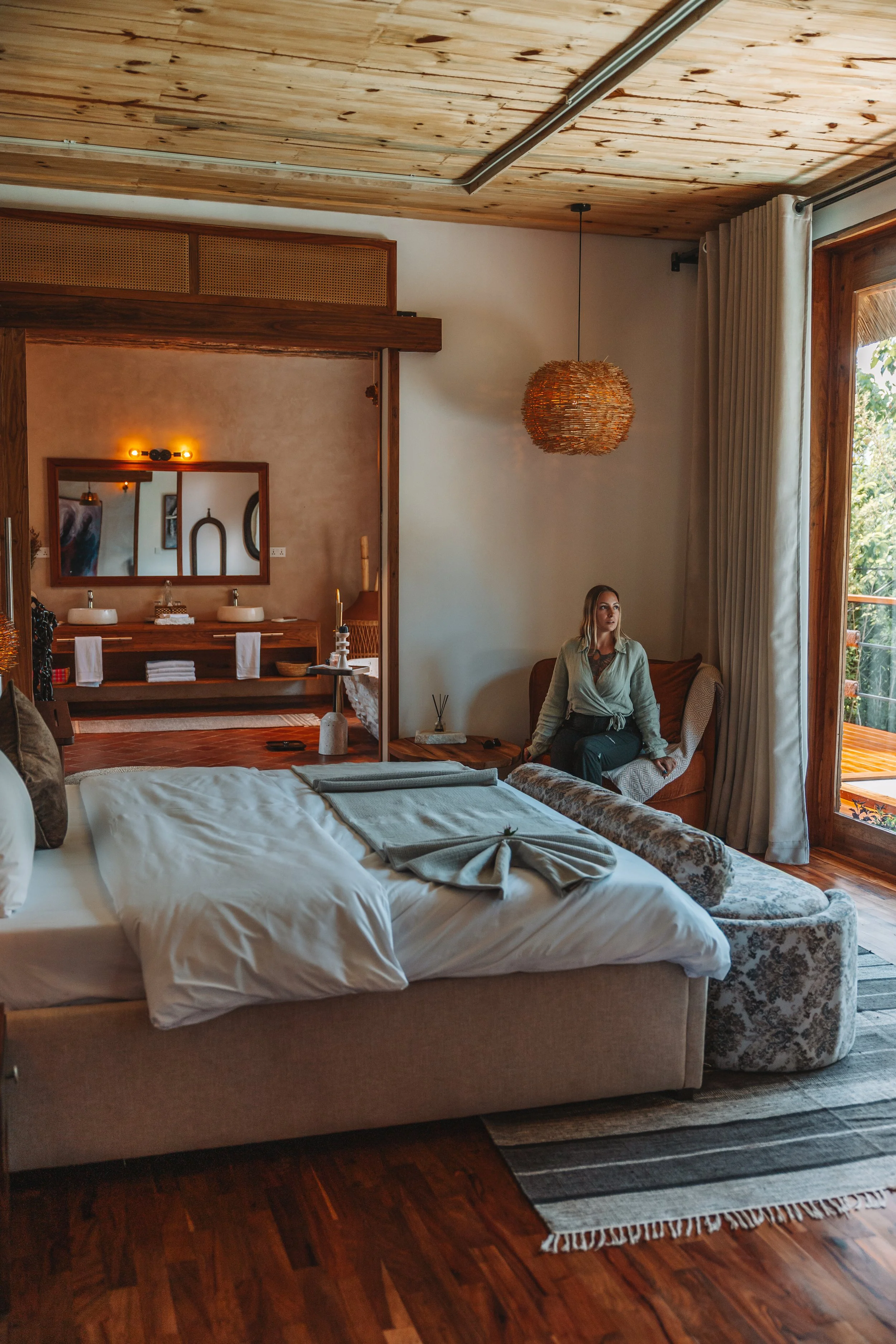 Interior of a cozy bedroom with a large bed, a woman sitting on a chair near a large window, a wooden ceiling, and warm decor including a pendant light and a striped rug.