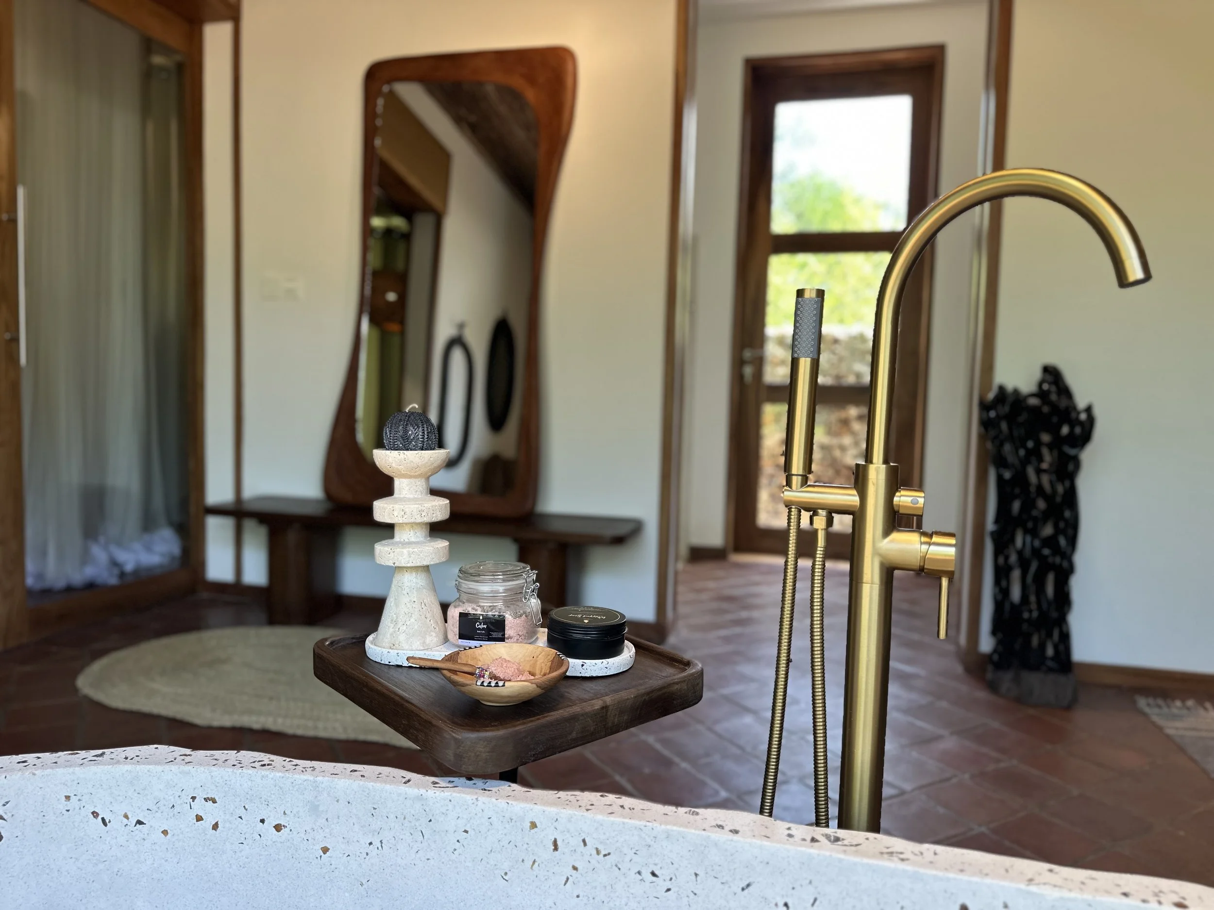 Interior of a house with a gold kitchen faucet, a wooden table with small containers, a decorative candlestick, a wooden bowl, and a mirror on the wall. Doors and windows showing greenery outside.