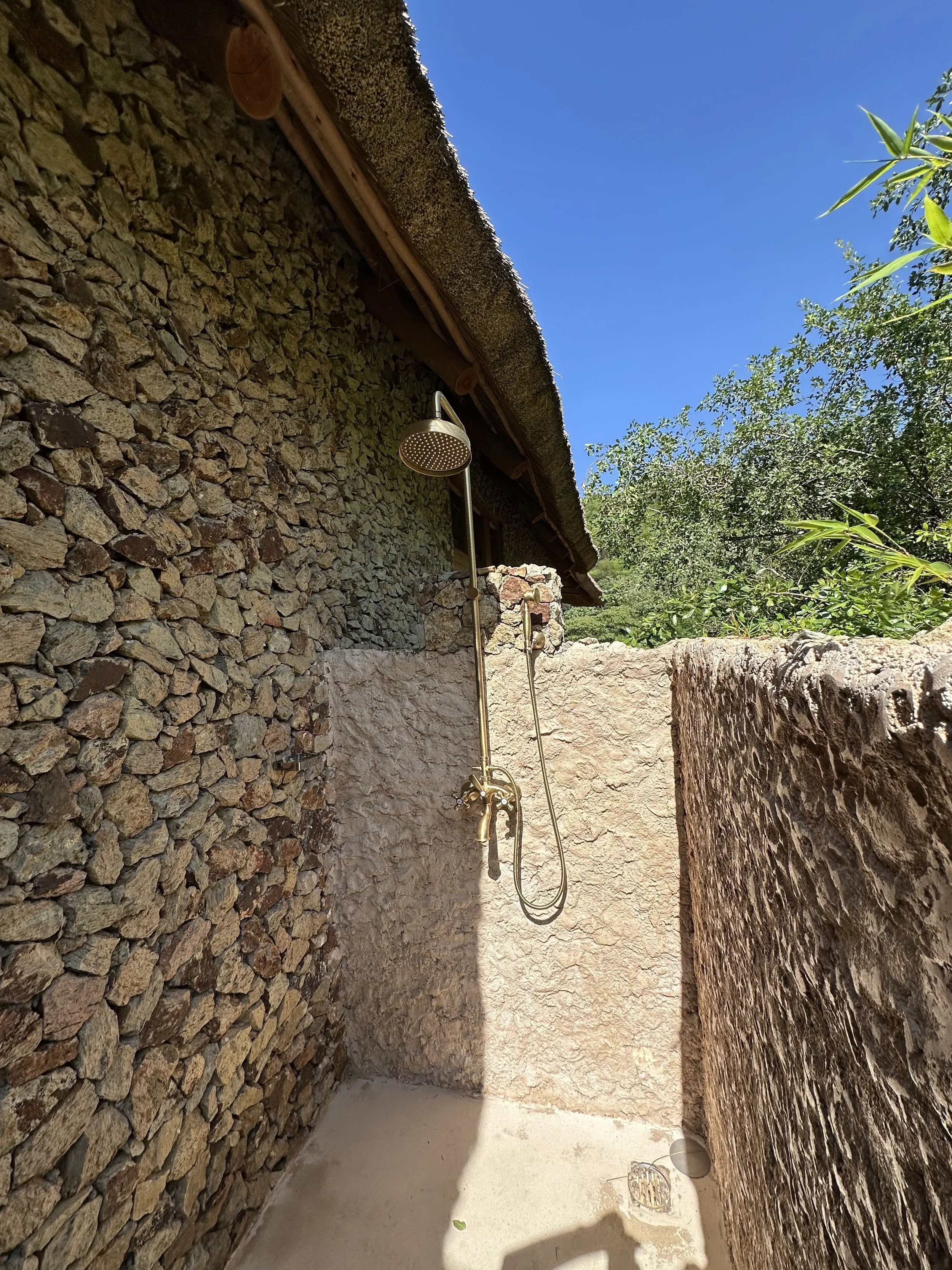 Outdoor shower area with a rustic stone wall and a rain shower head mounted on a wall, surrounded by green trees and a clear blue sky.