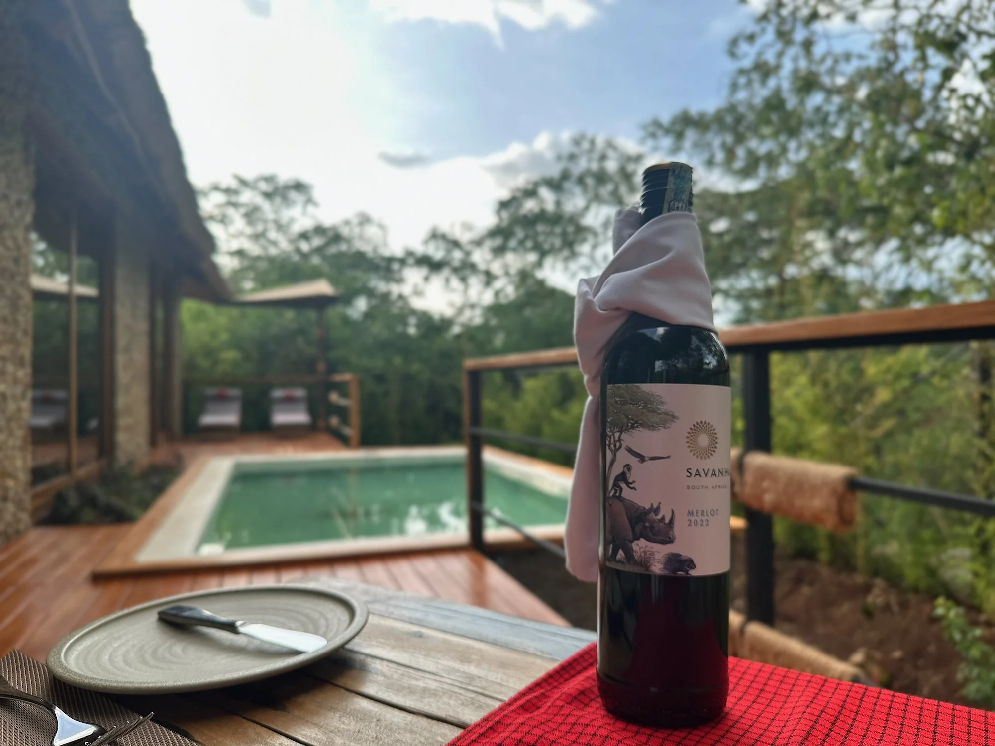 A bottle of red wine wrapped with a pink cloth, placed on a table with a red cloth, with a small knife on a plate nearby. In the background, there is a swimming pool, on a wooden deck, surrounded by trees and greenery, with a cloudy sky overhead.