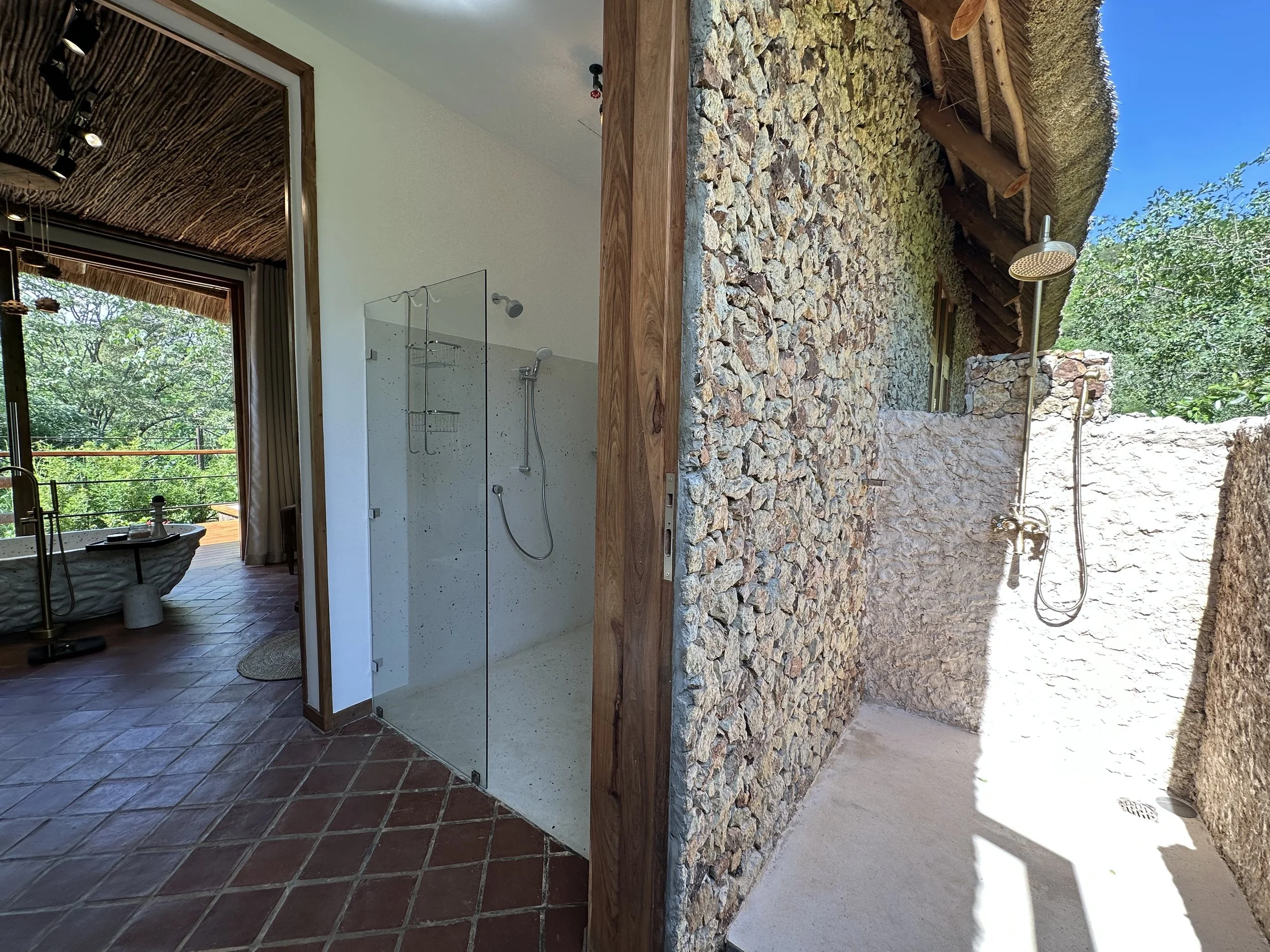 Split view of a bathroom with a walk-in shower and an outdoor shower area, surrounded by natural stone walls and lush greenery.