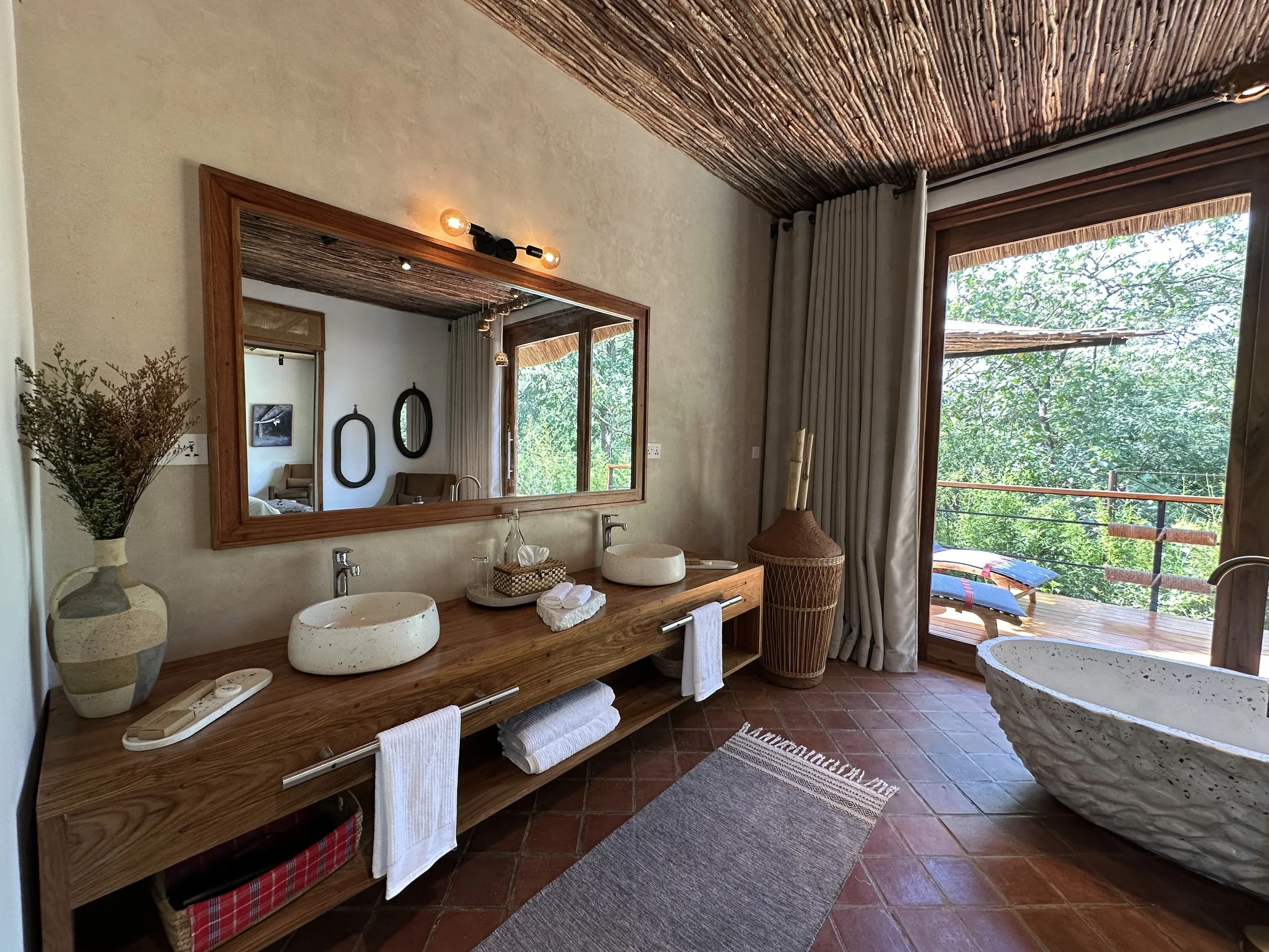 Luxury bathroom featuring a wooden double vanity with two stone sinks, a large wall mirror with wooden frame, a bathtub with a textured exterior, and a sliding glass door opening to a balcony with outdoor lounge chairs and lush greenery.