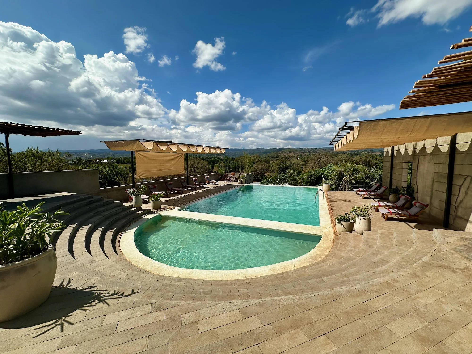 Outdoor swimming pool with lounge chairs and shade canopies, overlooking a scenic landscape of trees and hills under a partly cloudy sky.