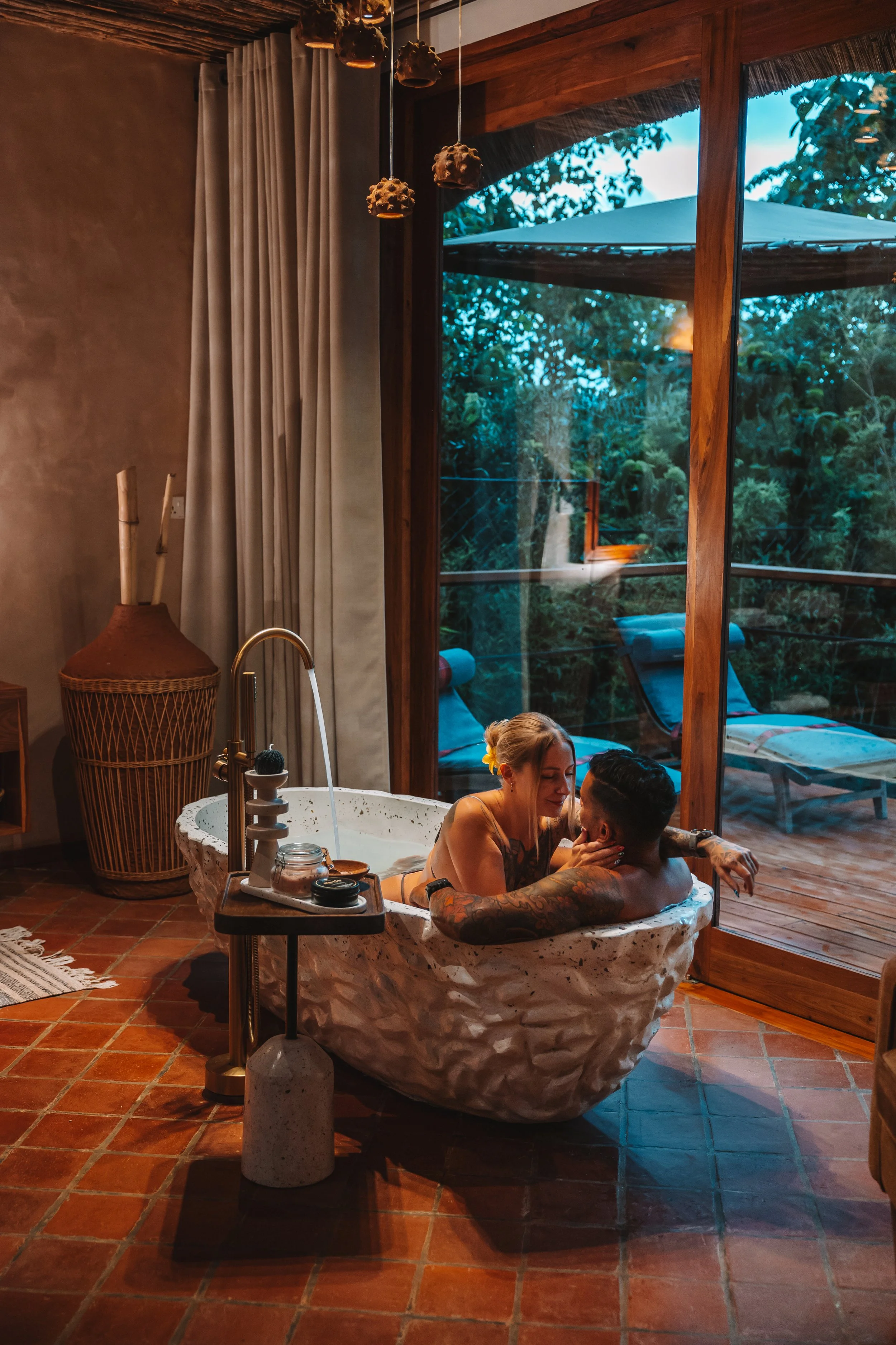 A couple enjoying a bath in a freestanding tub inside a cozy room with large window showing greenery and outdoor lounge chairs.