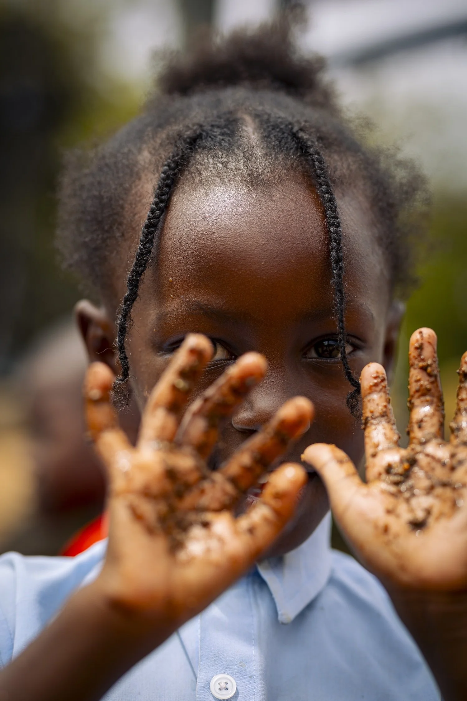 A young girl with braided hair, holding up her hands covered in mud or dirt, looking directly at the camera.