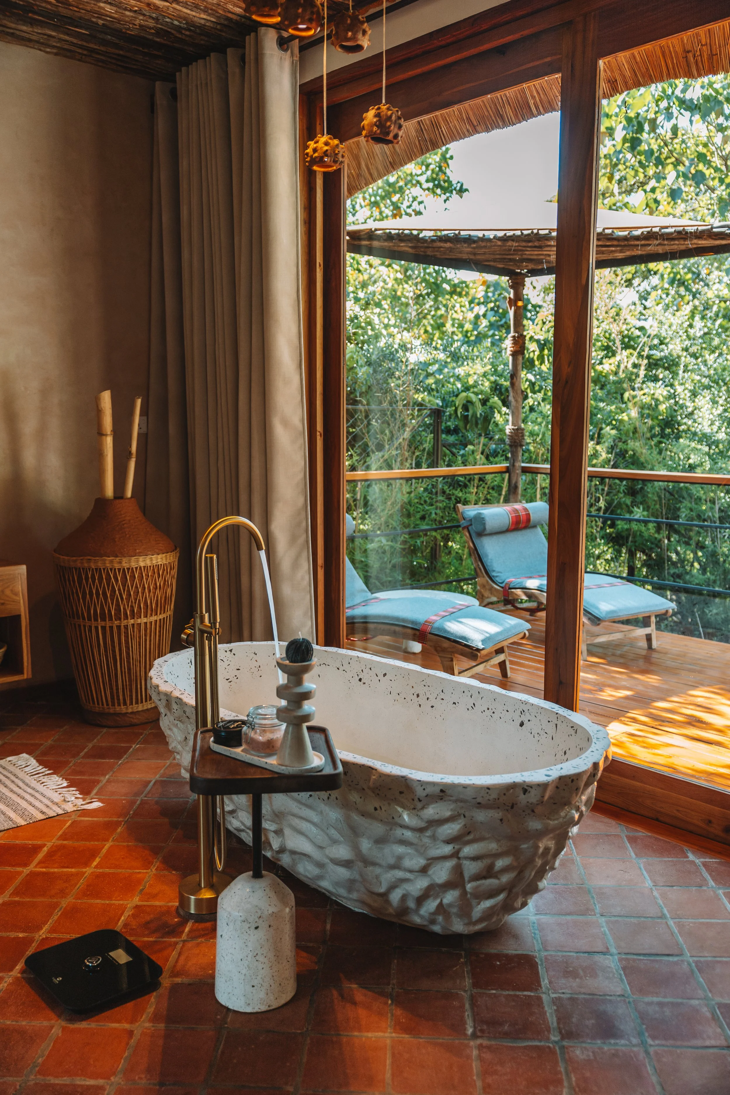 A bathroom with a large stone bathtub, a small tray holding personal care items, and a floor scale. There is a view of a wooden deck with lounge chairs outside, surrounded by green foliage.