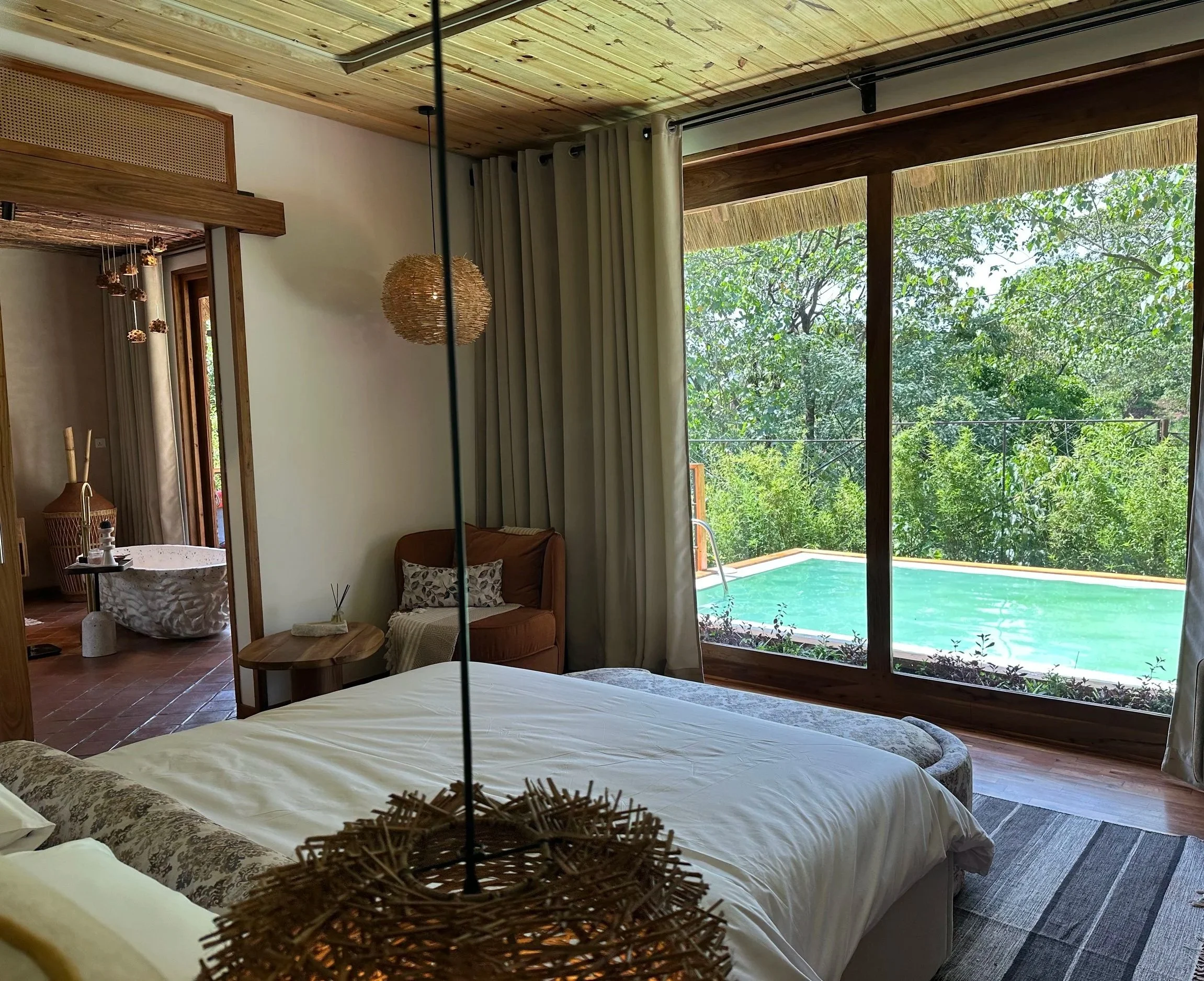 A cozy bedroom with a large window overlooking a swimming pool and lush green trees, featuring a bed, a small side table, an armchair, and a ceiling light fixture.