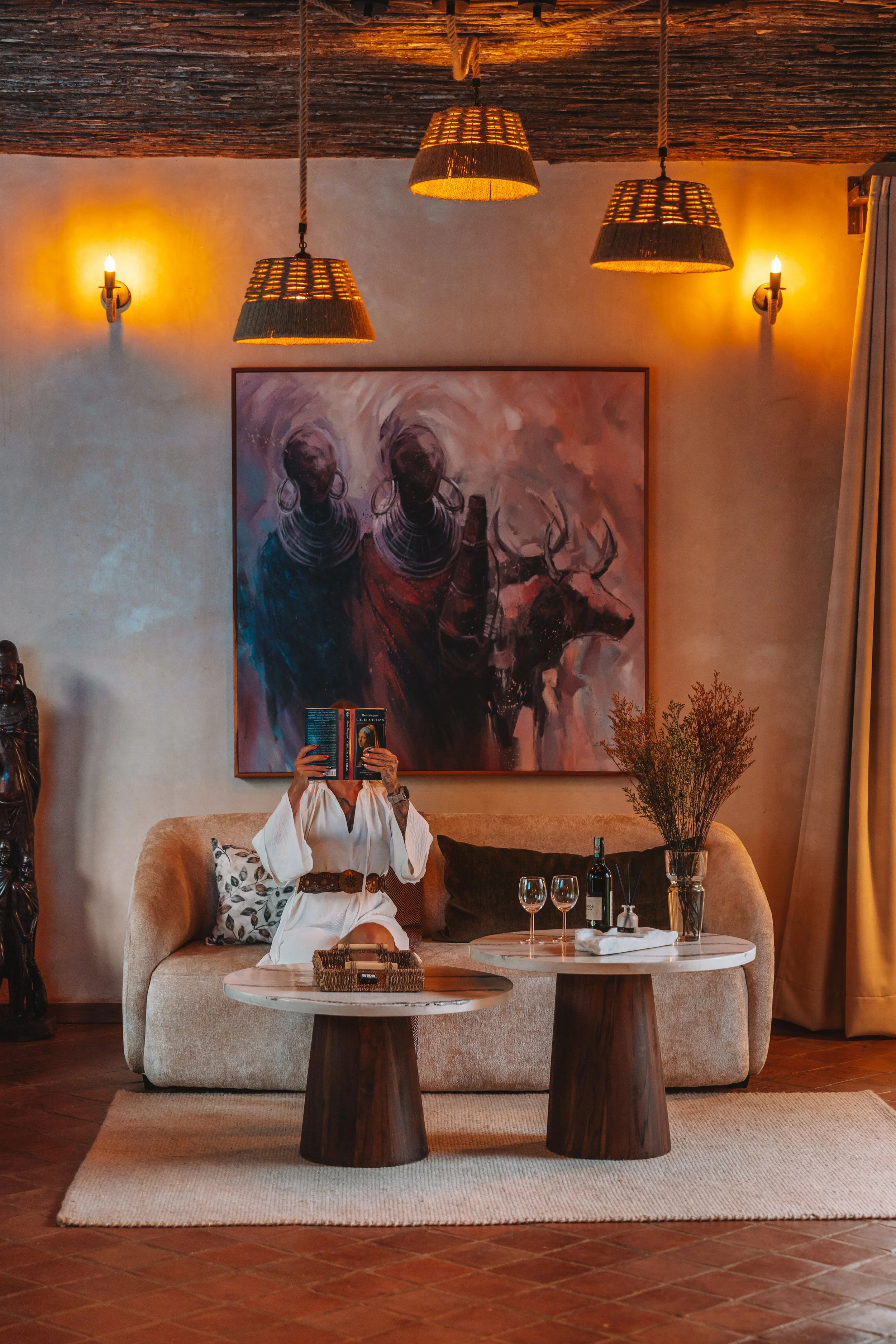 Interior of a cozy living room with a beige sofa, artwork of two women and a buffalo on the wall, wooden ceiling with hanging pendant lights, person sitting on sofa reading a book, two marble-topped tables with wine, glasses, and a plant, warm lighti