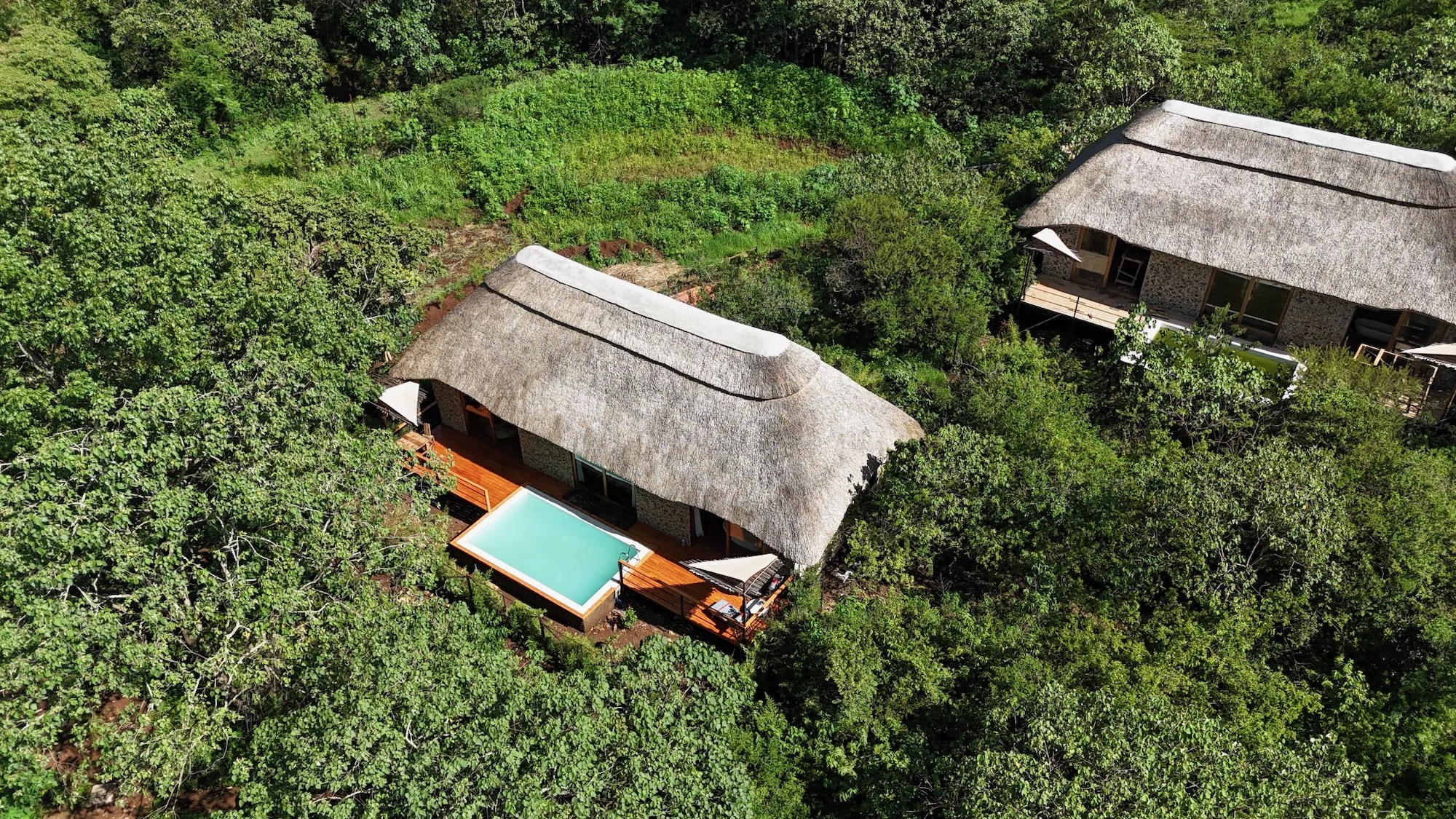 Aerial view of two thatched-roof houses surrounded by dense greenery, with a small pool and a wooden deck in front of one house.