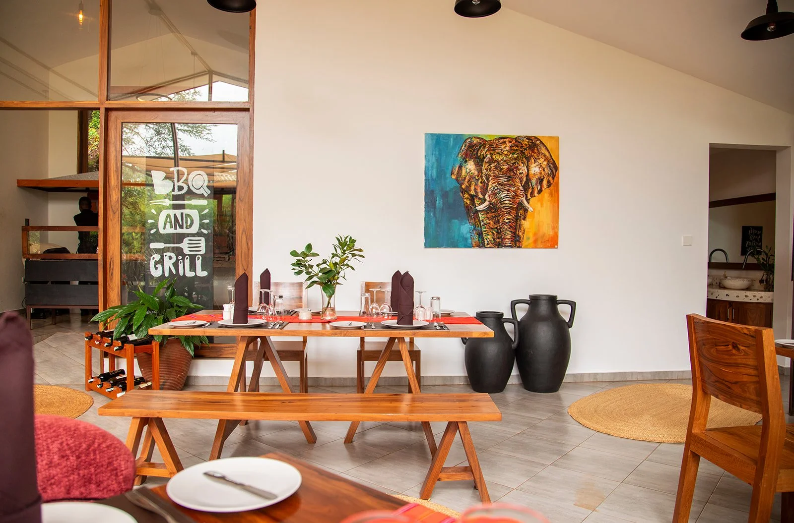 Interior of a restaurant dining area with wooden furniture, a colorful elephant painting on the wall, black ceramic vases, a large plant, and a window with a sign that says "BBQ AND GRILL."