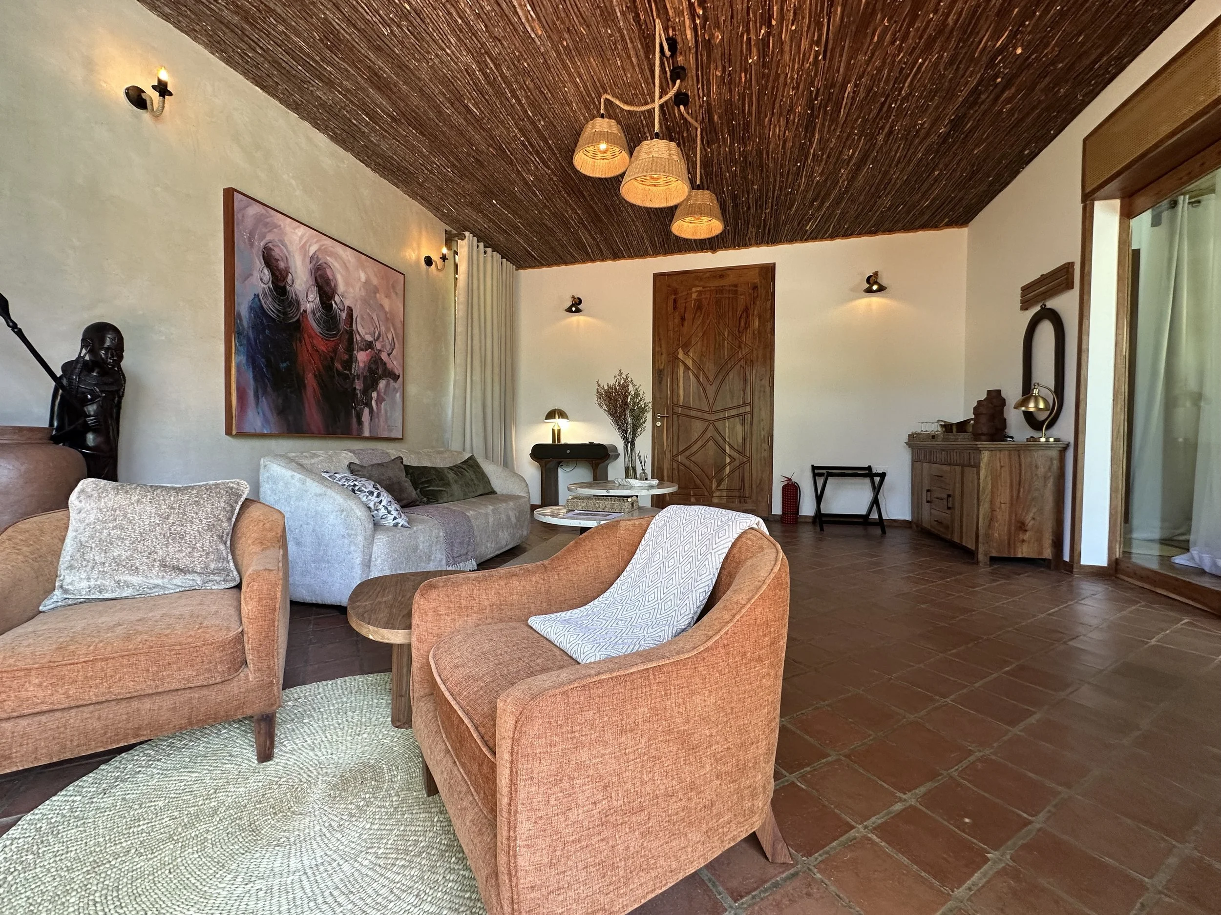 Living room with beige walls and a wooden ceiling, featuring two orange armchairs, a light gray sofa, a round coffee table, and wall-mounted lights. There's a large abstract painting on the wall, a wooden cabinet with decorative items, a small table 