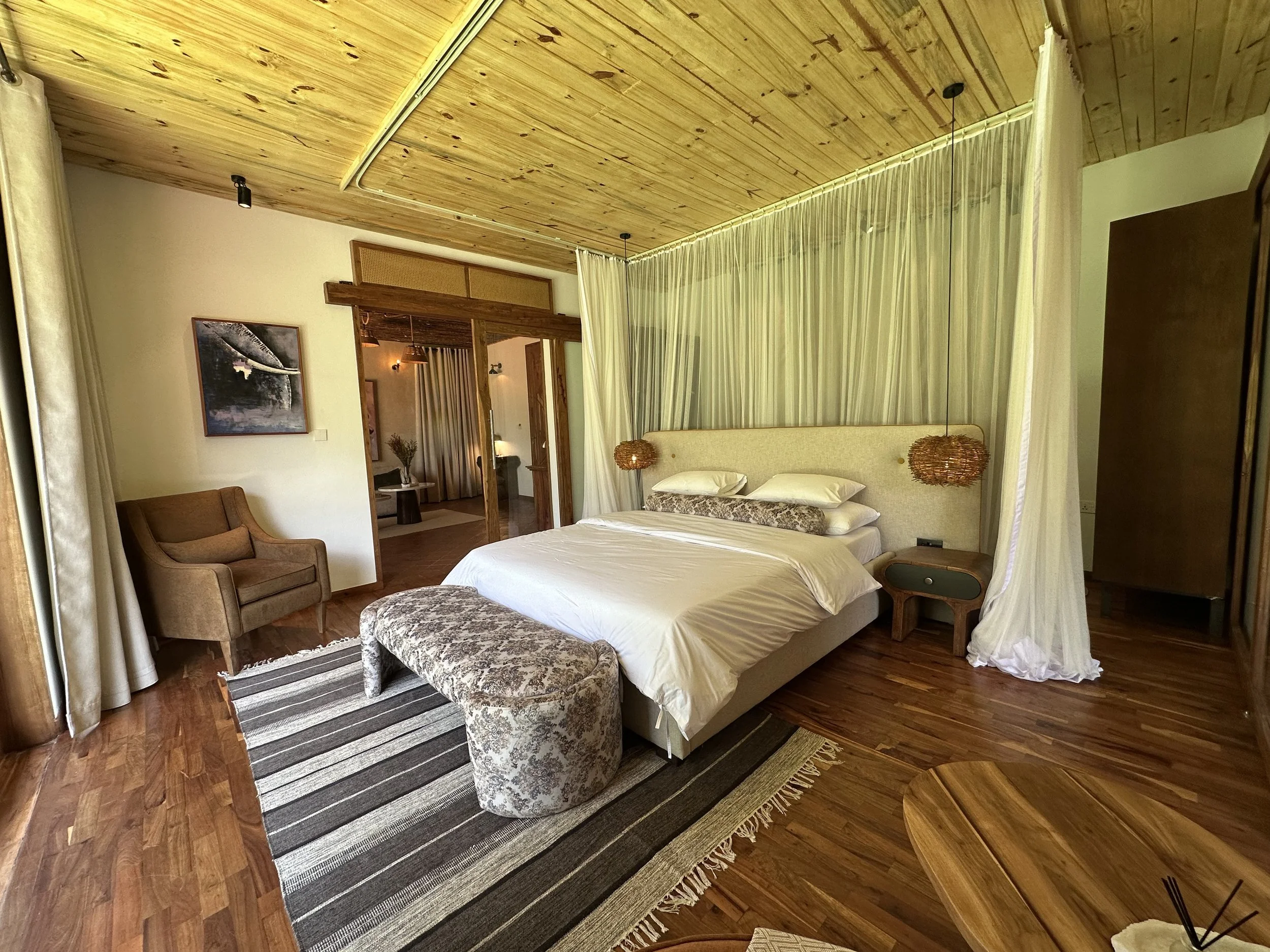 A hotel bedroom with a large bed, brown armchair, wooden ceiling, and sliding glass door leading to an adjoining room.